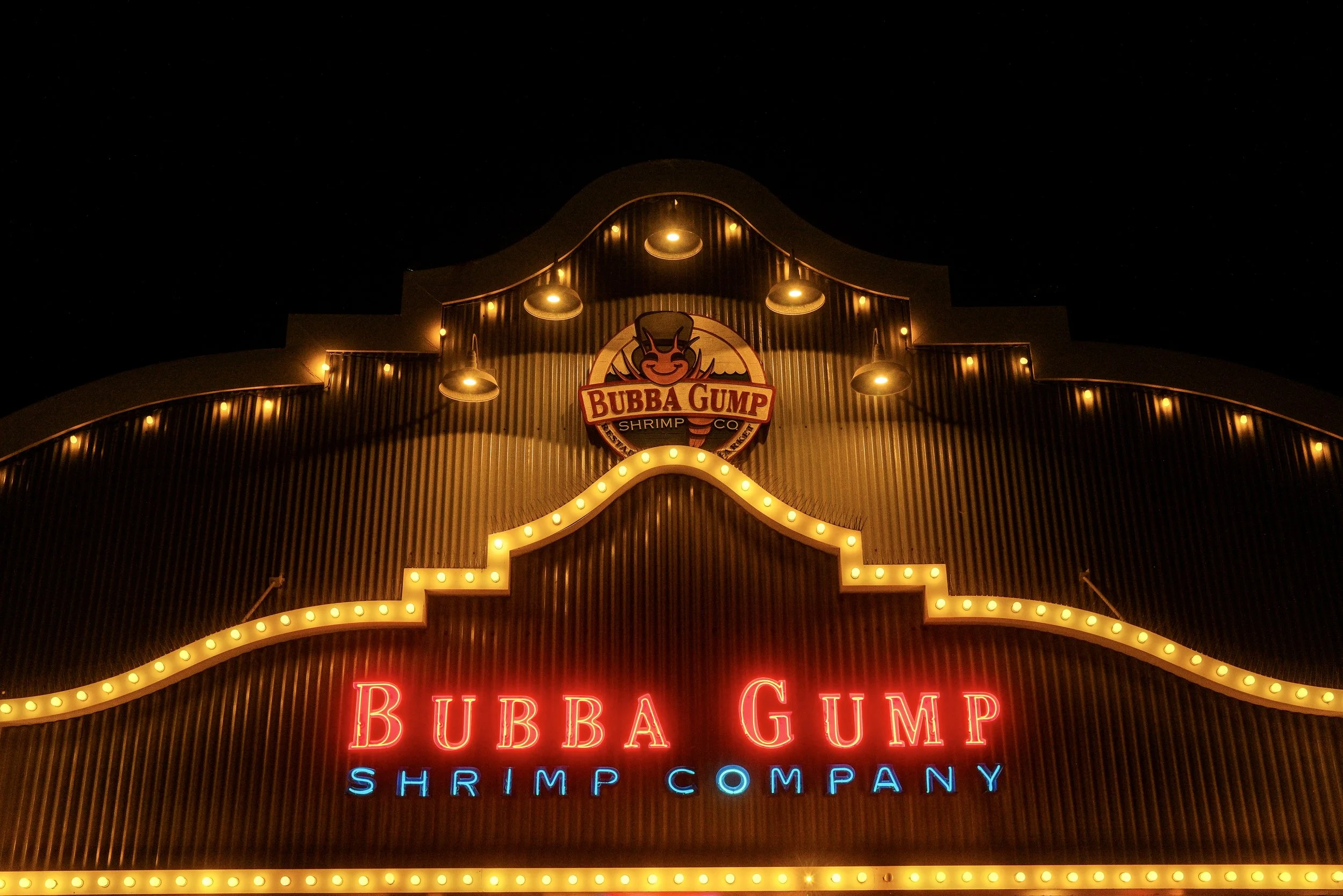 Night view of the illuminated exterior sign of Bubba Gump Shrimp Company restaurant with bright red and blue neon lights, featuring the restaurant's logo at the top center.