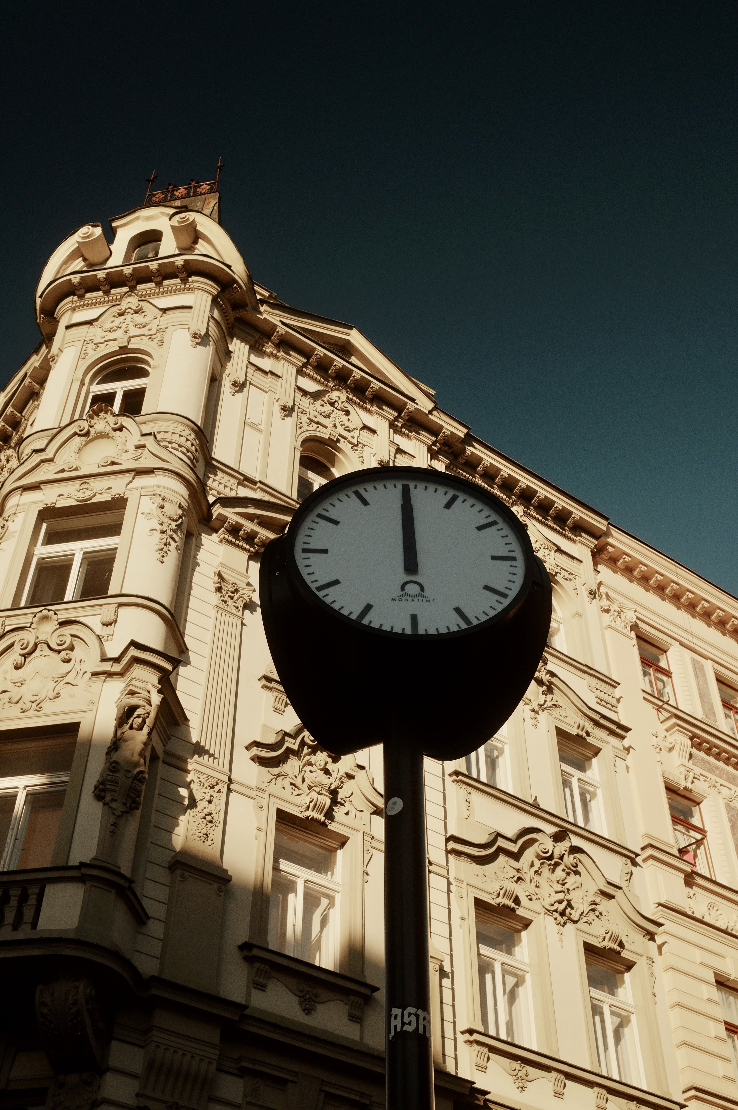 A street clock showing the time of 12:00, with an ornate historic building in the background.