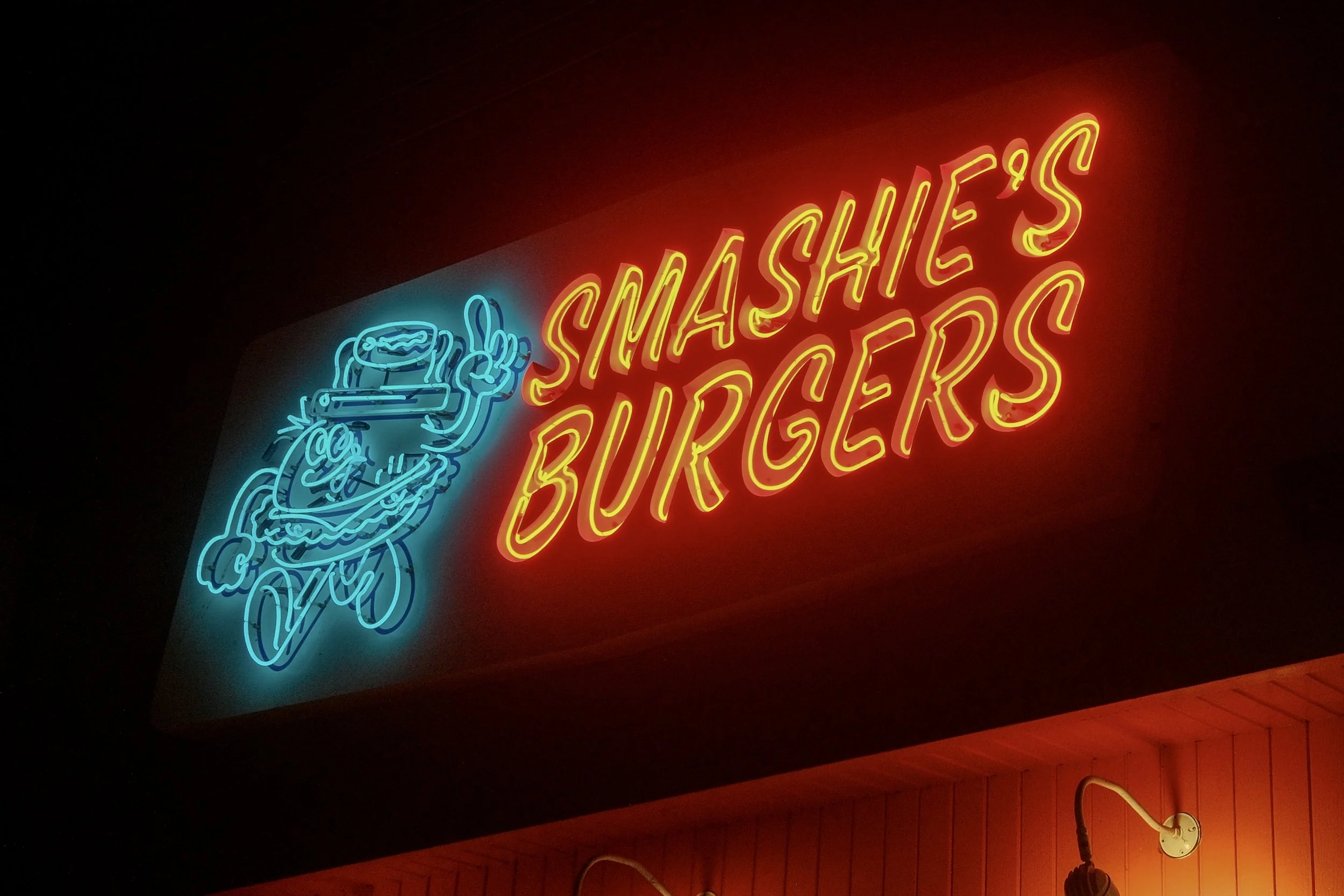 Neon sign with a cartoon burger character giving a peace sign next to the words 'Smashie's Burgers' in bright red and yellow letters, against a dark background.