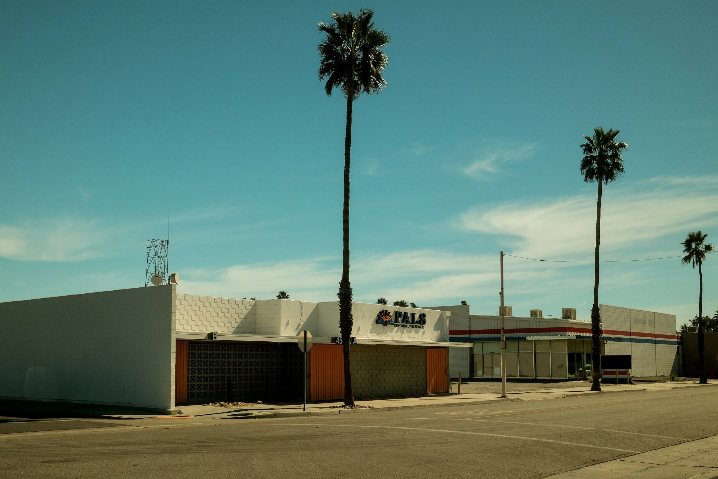 A single-story commercial building under a clear blue sky with tall palm trees in front. The building has a sign that reads 'PALS Supported Living Services.'