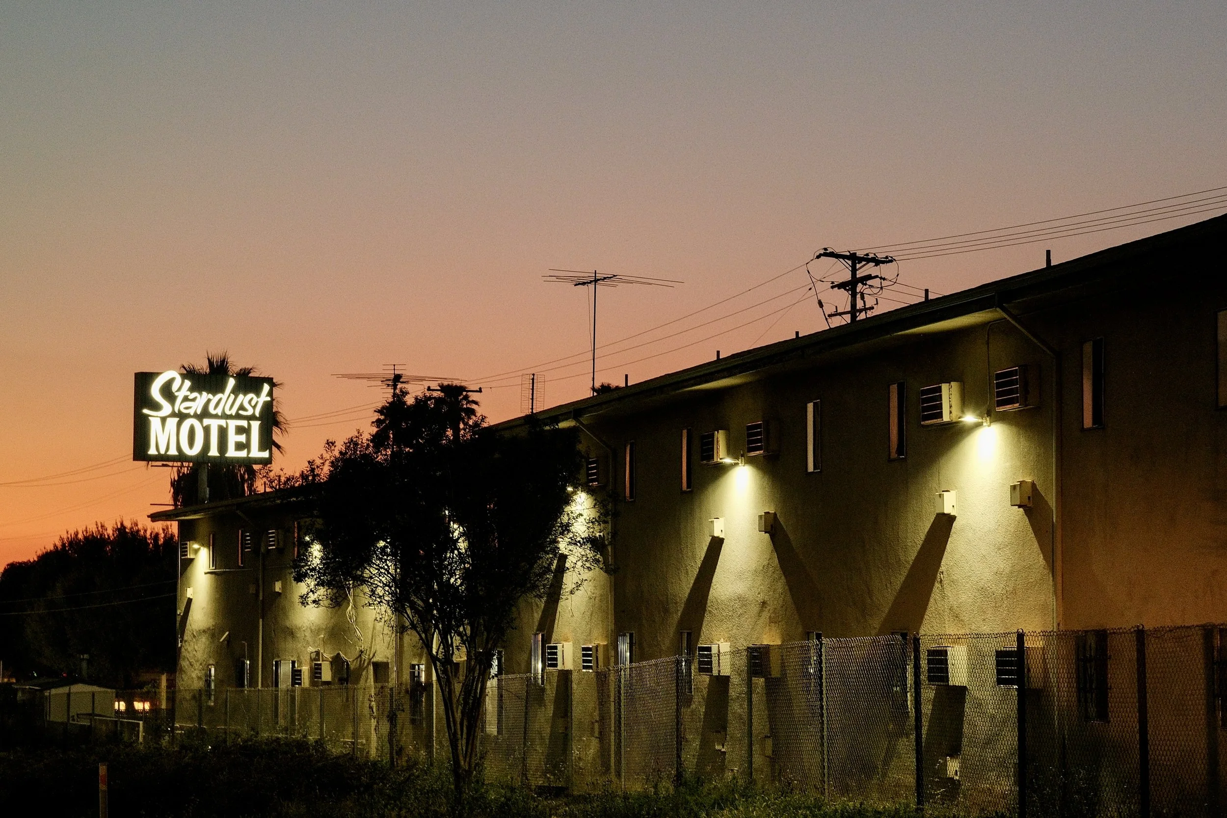 The exterior of the Stardust Motel at dusk with a lit sign, trees, a fence, and a building with small windows and wall-mounted lights, against a sunset sky.