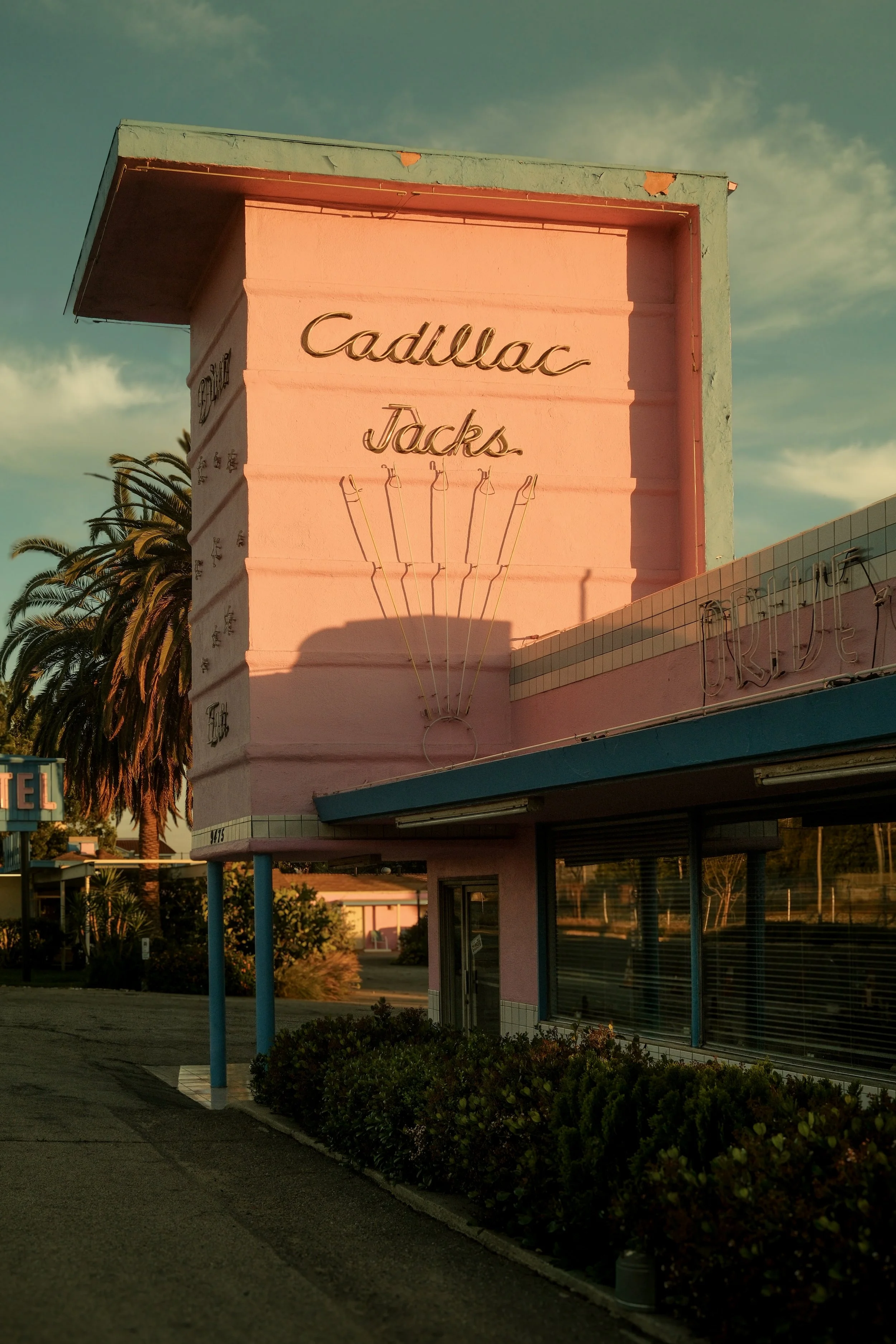 The exterior of a pink vintage building with a large sign that reads 'Cadillac Jacks' in cursive, illuminated by warm sunlight, with palm trees in the background.