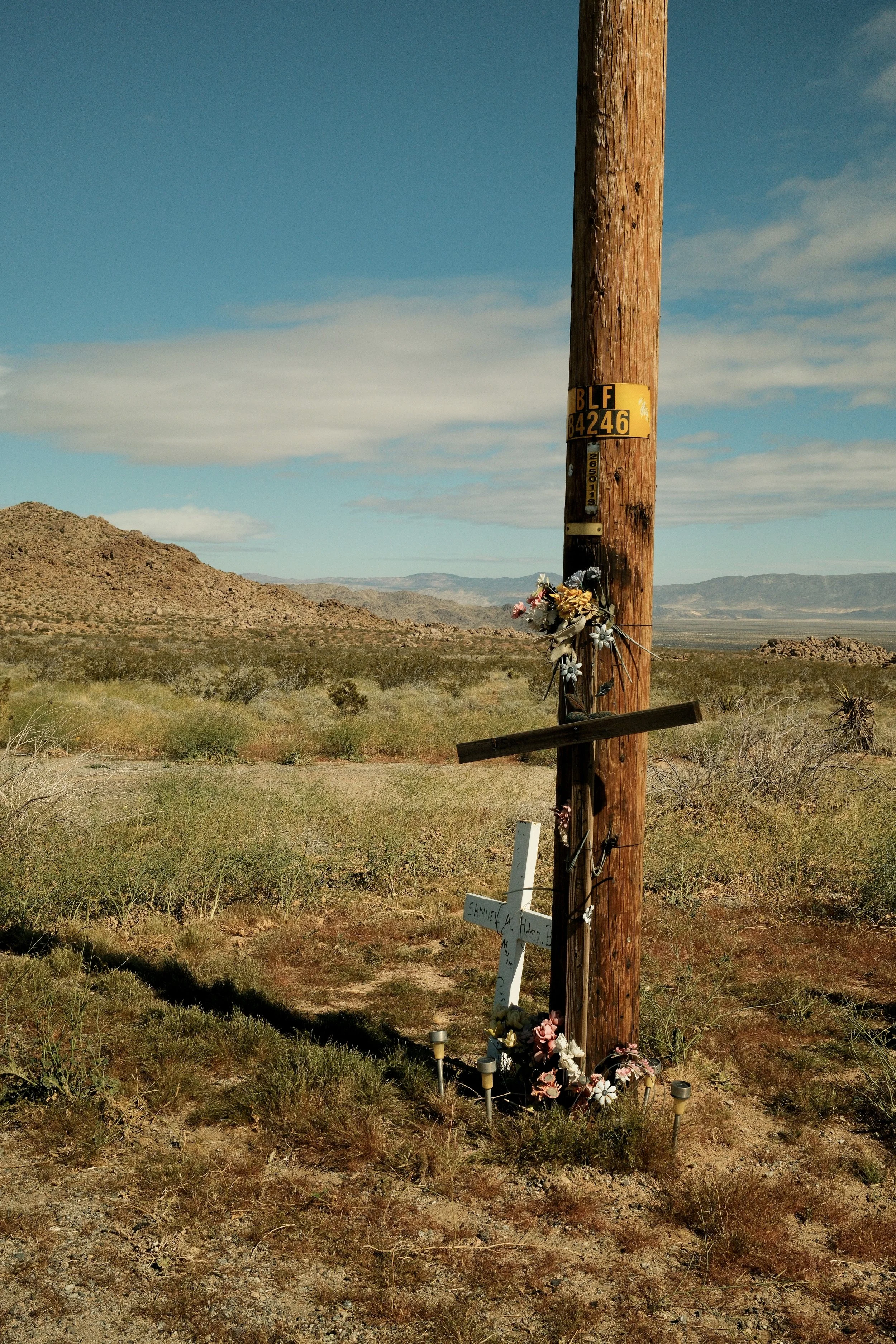 A wooden utility pole with flowers and a cross at its base, set in a desert landscape with mountains in the background.