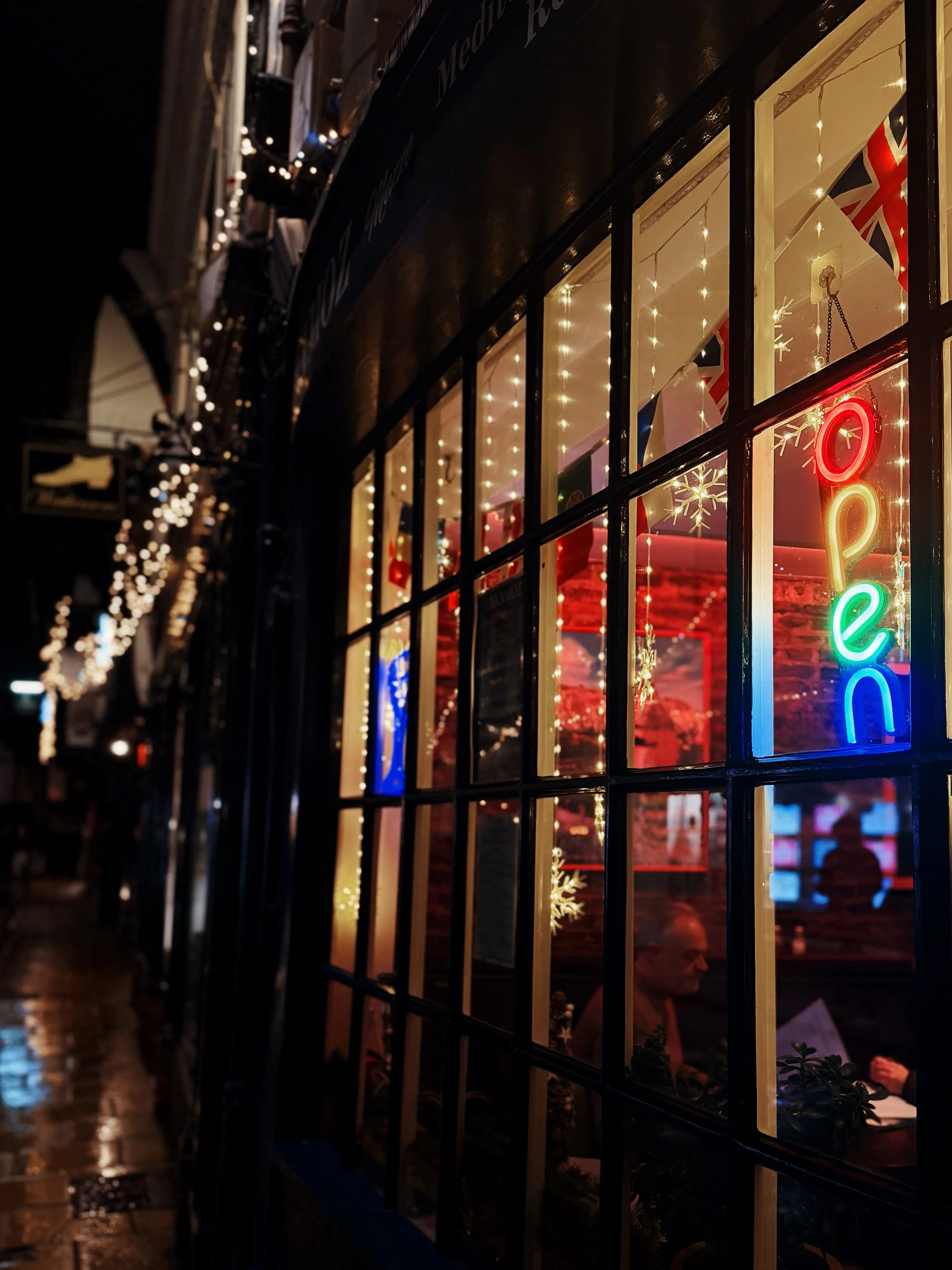 Nighttime view of a restaurant window decorated with string lights and a colorful neon door sign that says "open". Inside, a person is sitting at a table with a menu. Outside, the sidewalk and city street are visible.