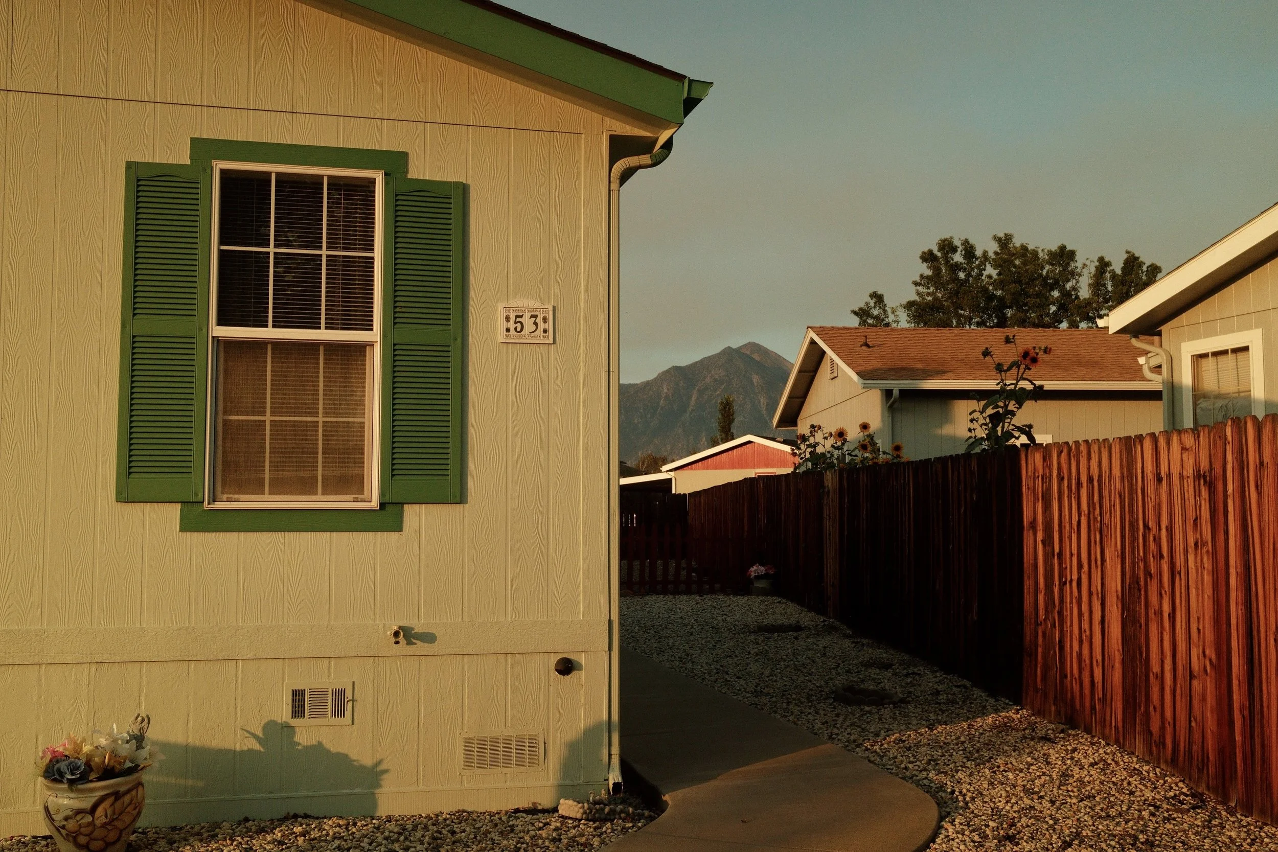 Exterior view of a house with a green window shutter, a house number 53, a garden corner with flowers, and a pathway leading to other houses, with mountains in the background at sunset.