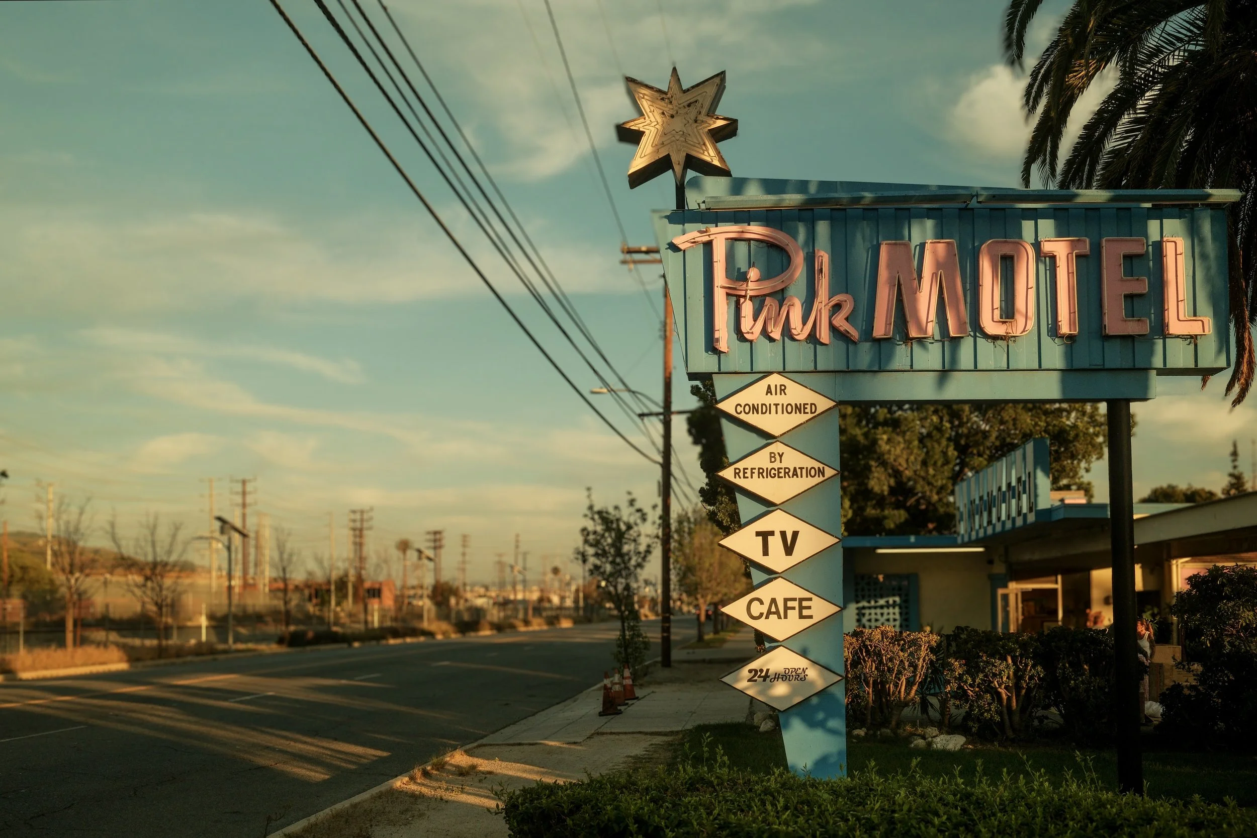 A vintage neon sign for Pink Motel with additional signs indicating air-conditioned rooms by refrigeration, TV, cafe, and 24-hour operation, in a small town setting during sunset.