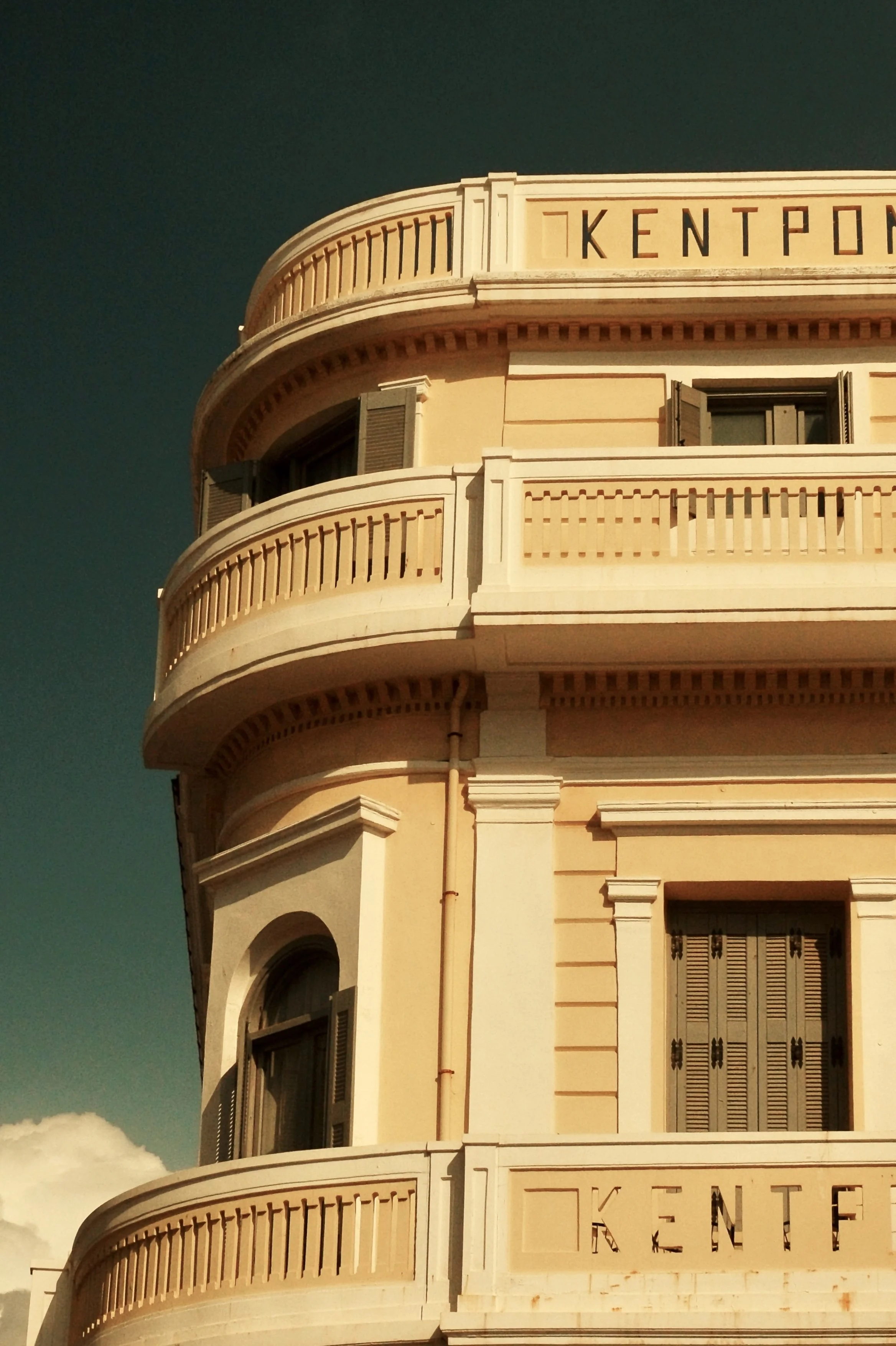 Corner view of a classical building with cream-colored stone, rounded balconies with decorative railings, and windows with dark shutters, with the word 'KENT' partially visible at the top and bottom.