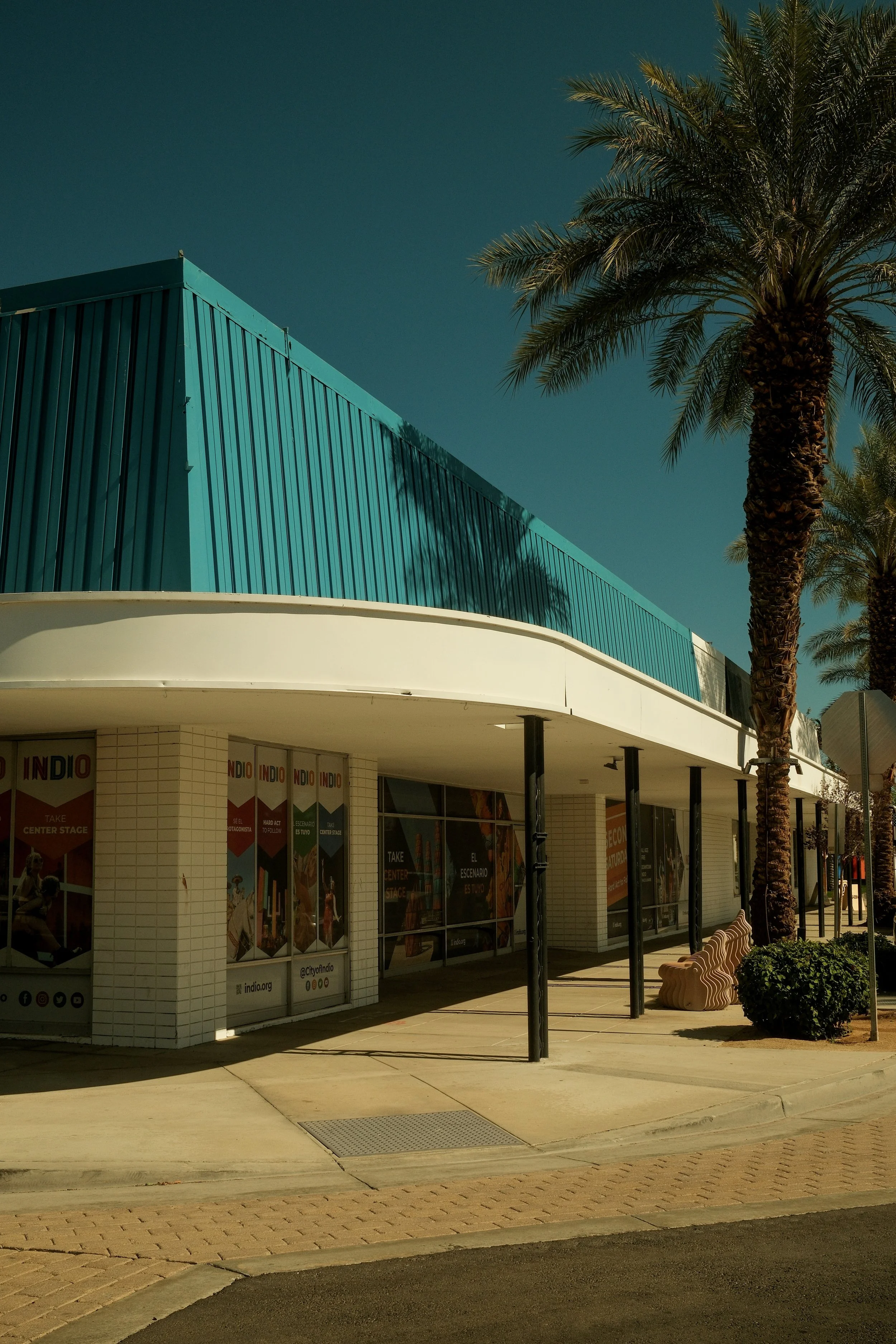 A modern building with teal metal siding, large windows, and white walls, surrounded by tall palm trees on a sunny day.