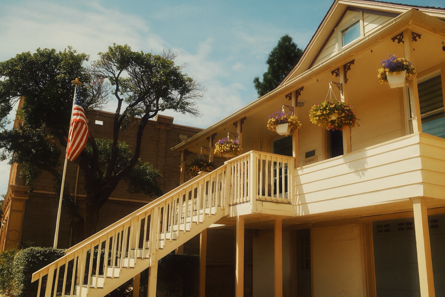 A yellow house with hanging flower baskets and a white staircase leading to the front door, an American flag on a flagpole, and large trees in the background under a partly cloudy sky.