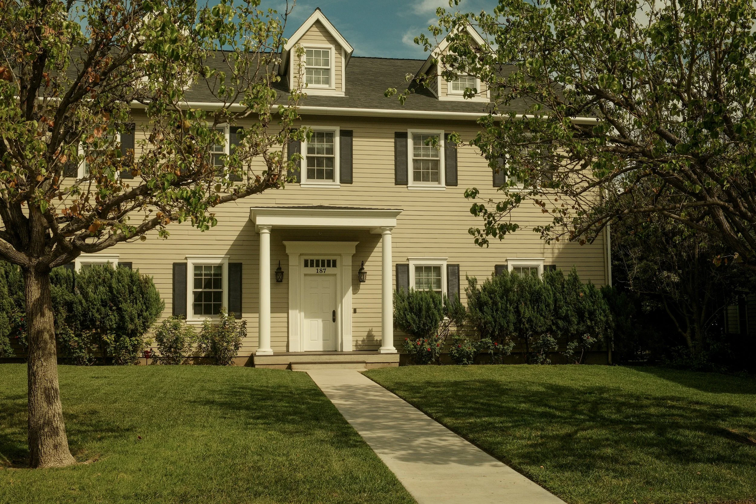 A beige, multi-story house with black shutters, a white front door, and a porch supported by two columns. The house number is 187. There are two trees with green leaves framing the house, and a green lawn with a sidewalk leading to the front door.