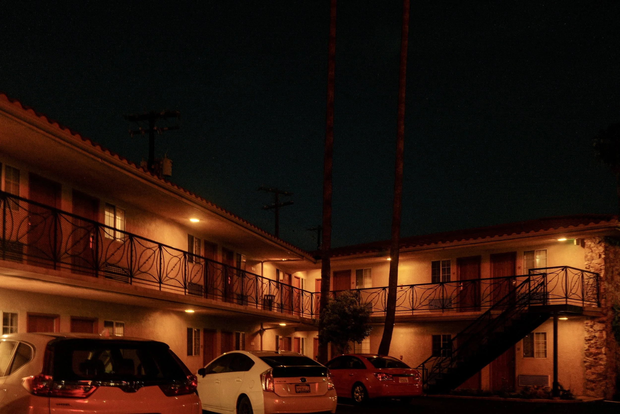 Exterior view of a motel courtyard at night with lit windows, fire escape stairs, parked cars, and palm trees.