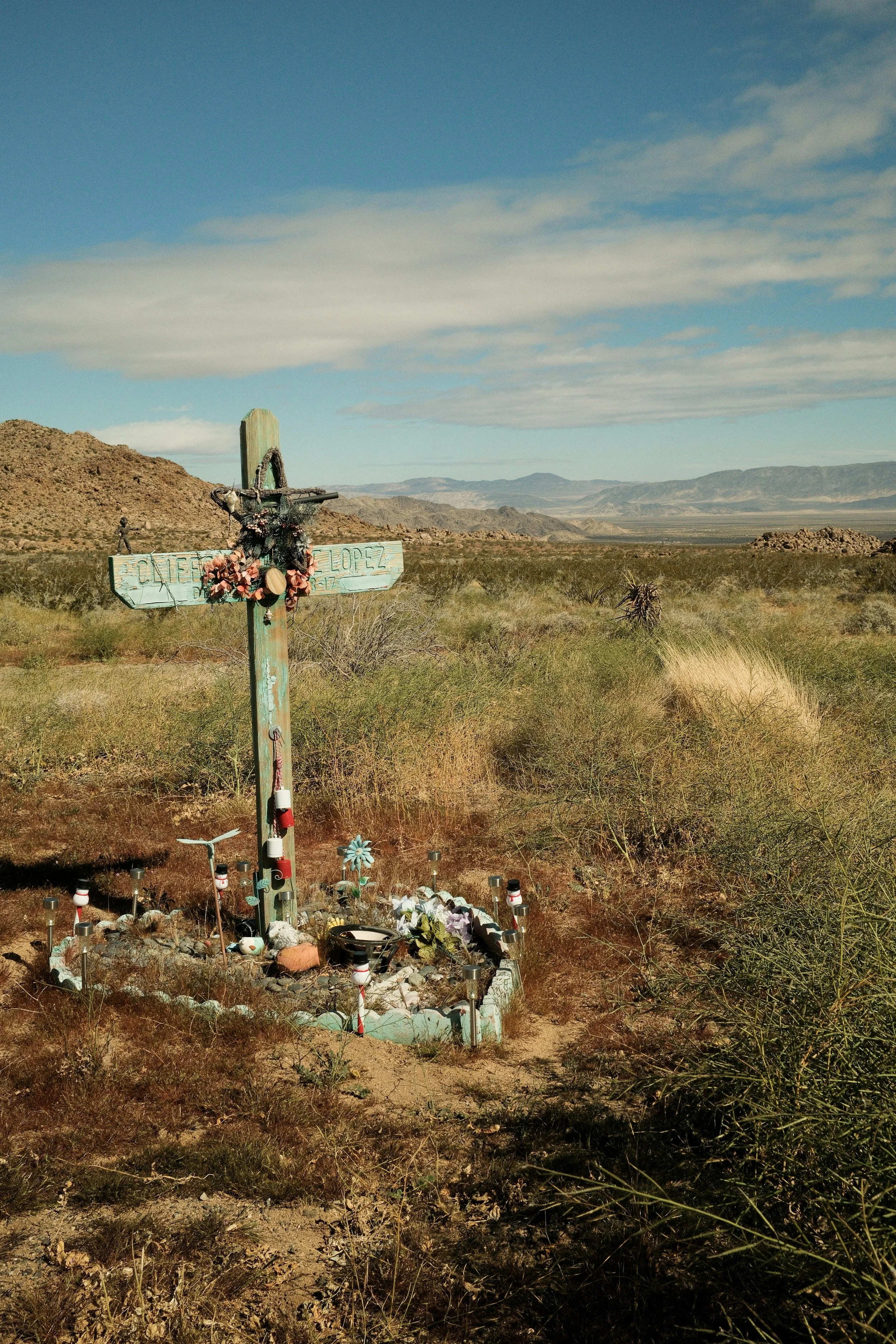 A small roadside memorial with a cross decorated with flowers and ribbons, surrounded by small decorative items on a desert landscape with mountains in the background.