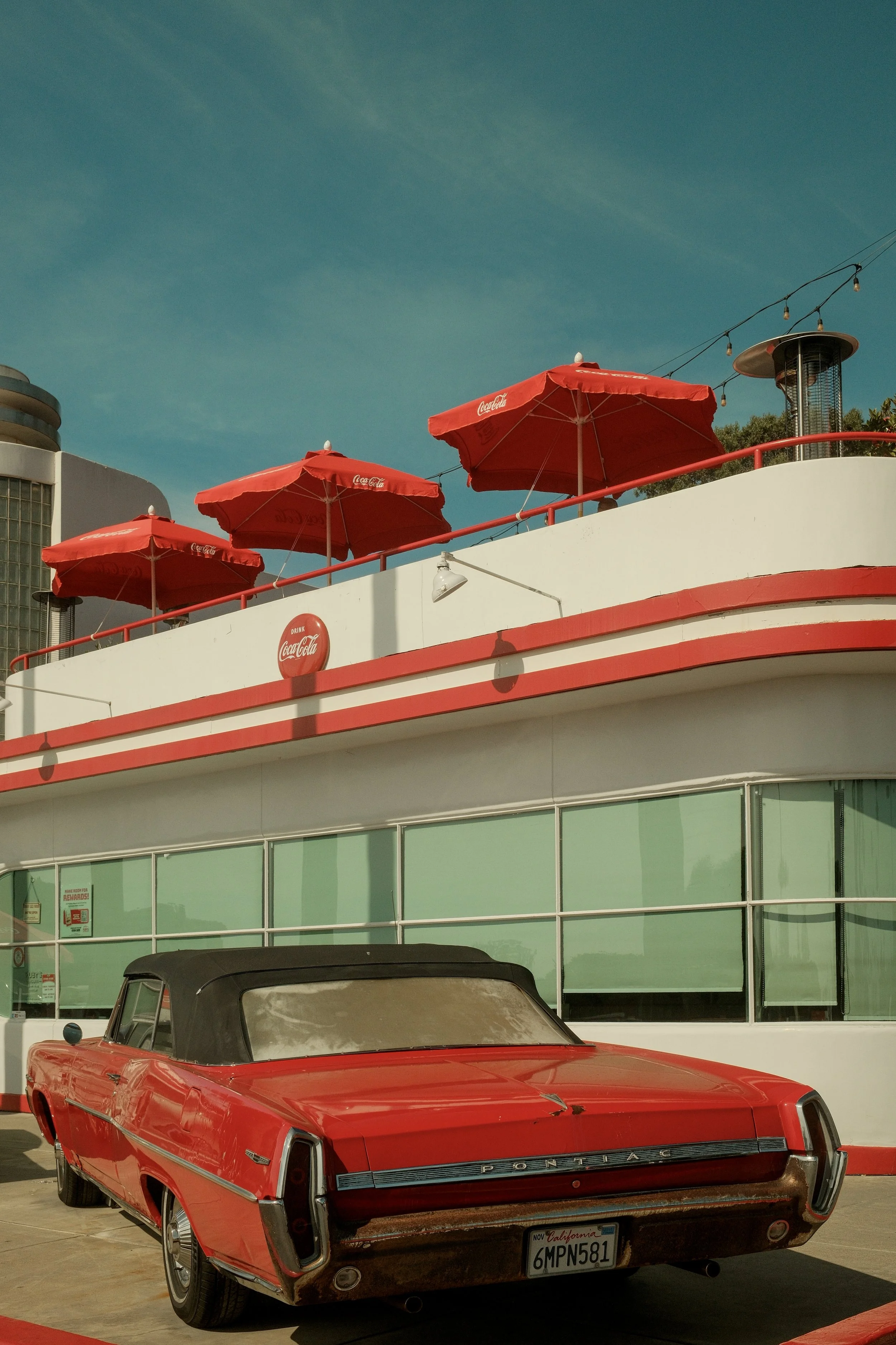 Vintage red Pontiac car parked outside a retro diner with a white facade, large windows, and red accents. The diner has three red Coca-Cola umbrellas on the rooftop.
