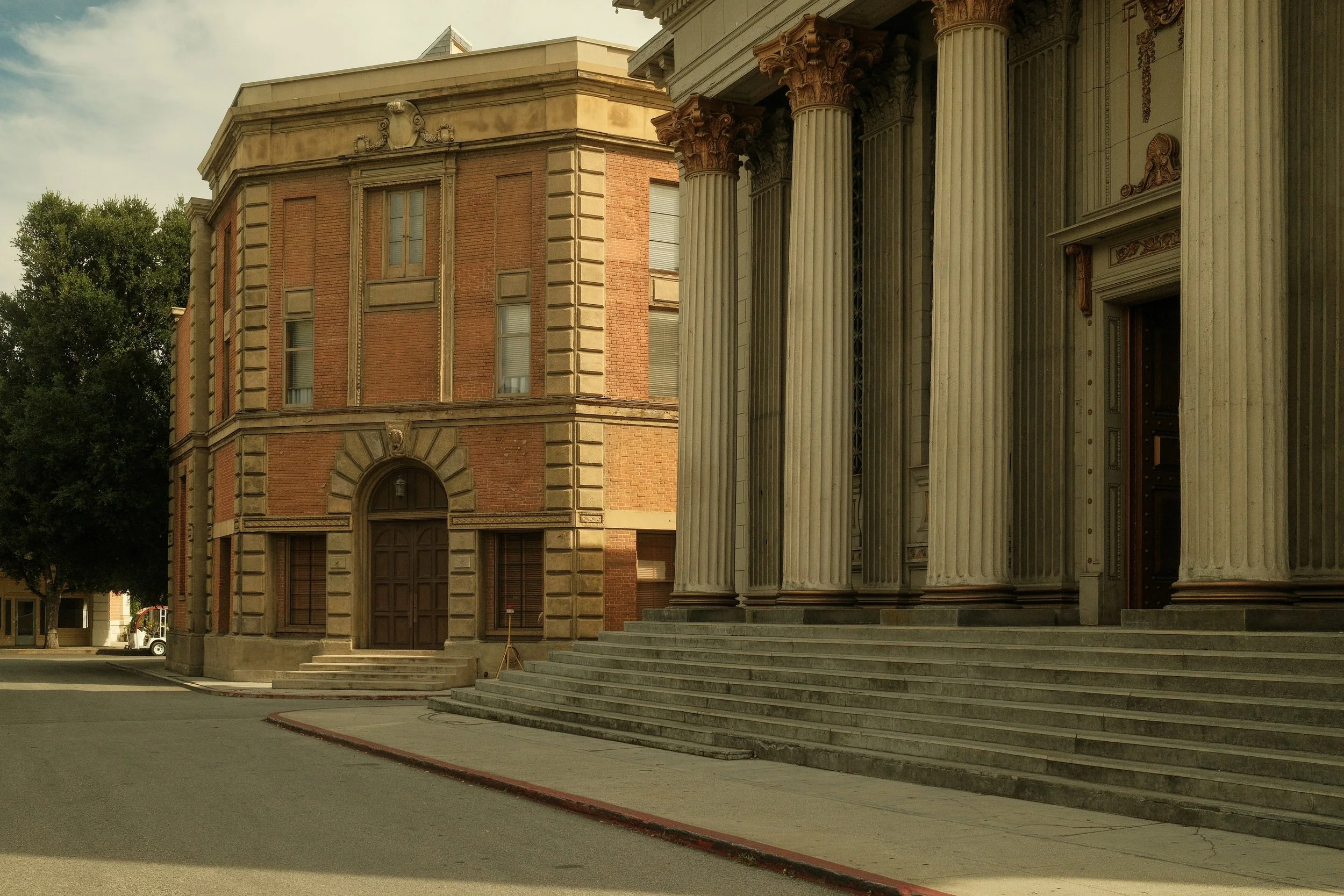 An old building with classical architecture, tall columns, and steps leading to an entrance, located next to a shorter brick building on a quiet street.