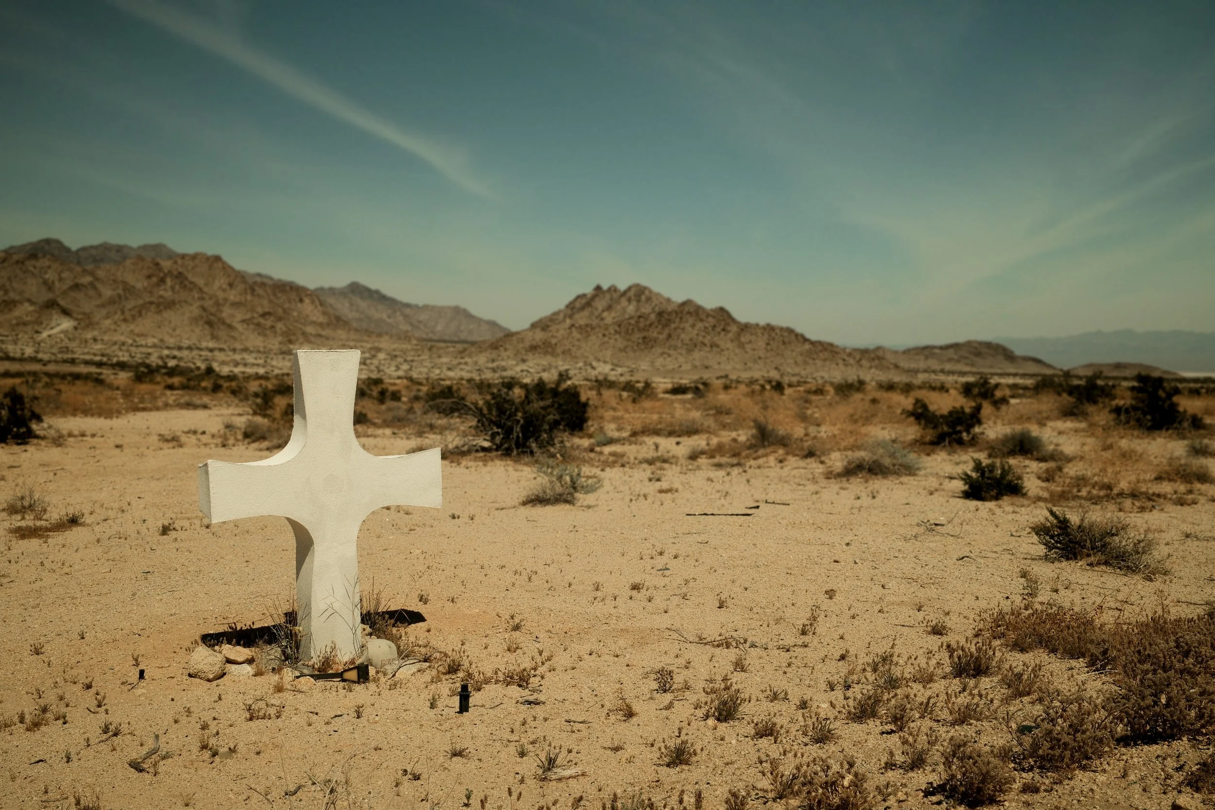 White cross memorial in a desert landscape with mountains in the background and sparse bushes under a blue sky.