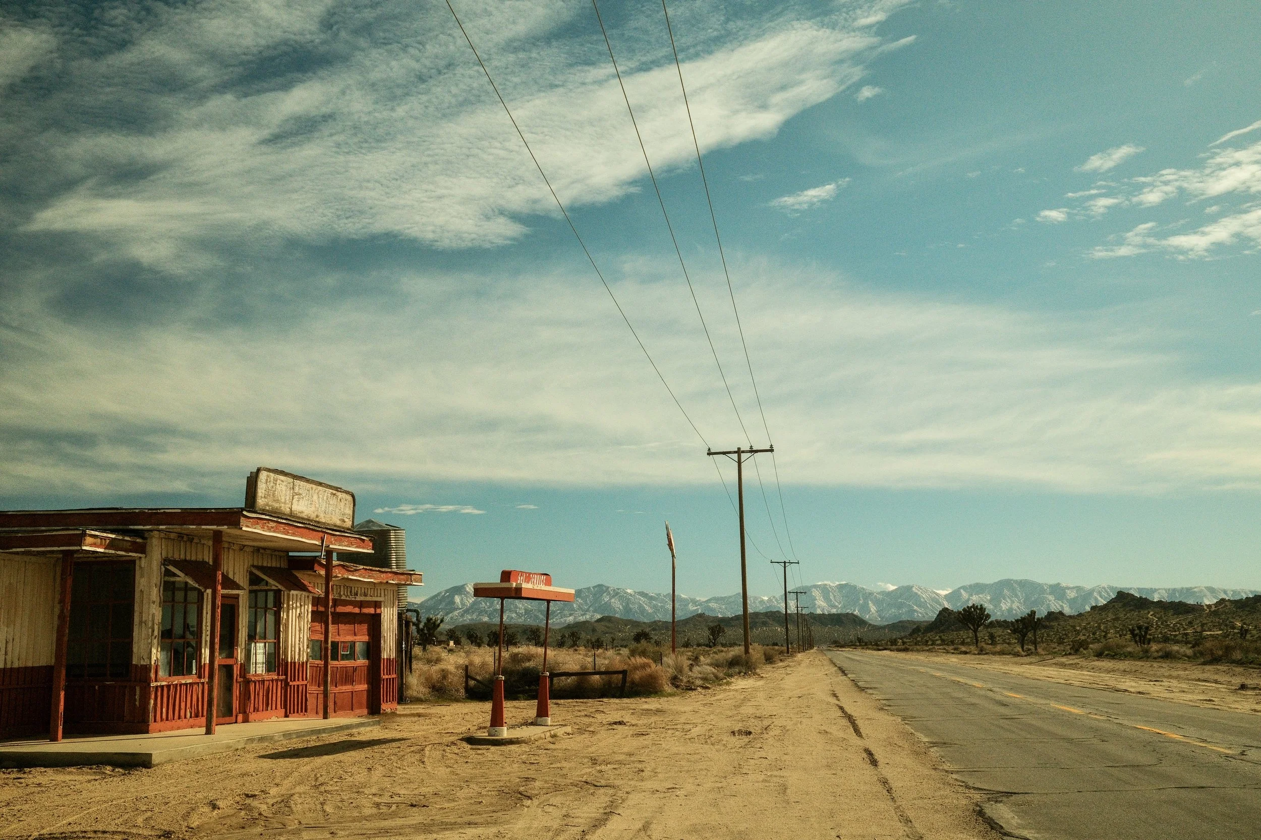 A deserted roadside with an old, weathered building on the left and a dirt shoulder on the right. Utility poles with wires extend into the distance, and distant snow-capped mountains are visible under a partly cloudy sky.