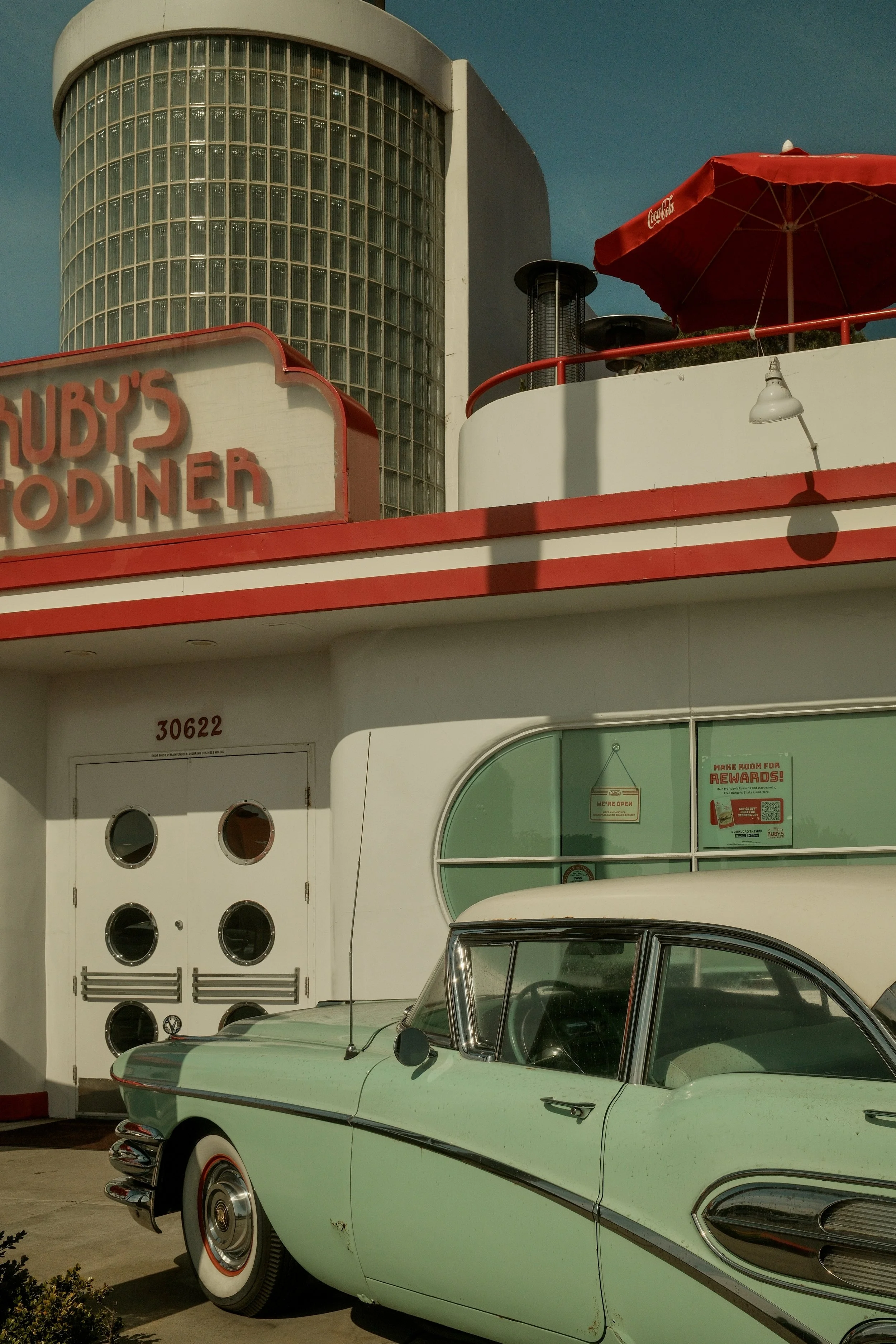 A vintage green and white car parked outside a building with a Mid-Century Modern diner sign that reads 'Ruby's Diner', with a curved facade and large glass block window on the upper level, and a red Coca-Cola umbrella on a balcony.