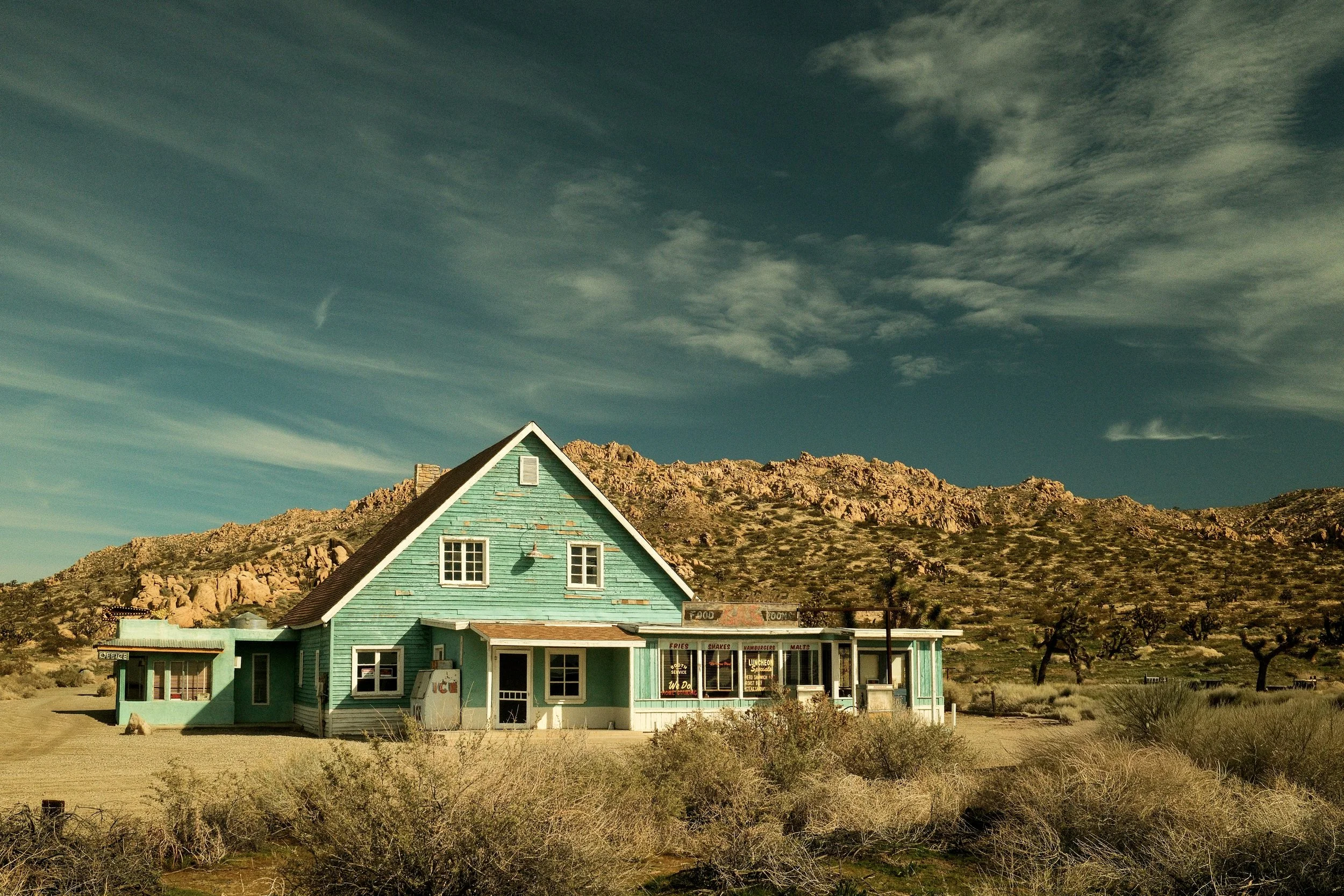 A turquoise wooden house in a desert landscape with sparse bushes and rocky hills in the background under a blue sky with scattered clouds.