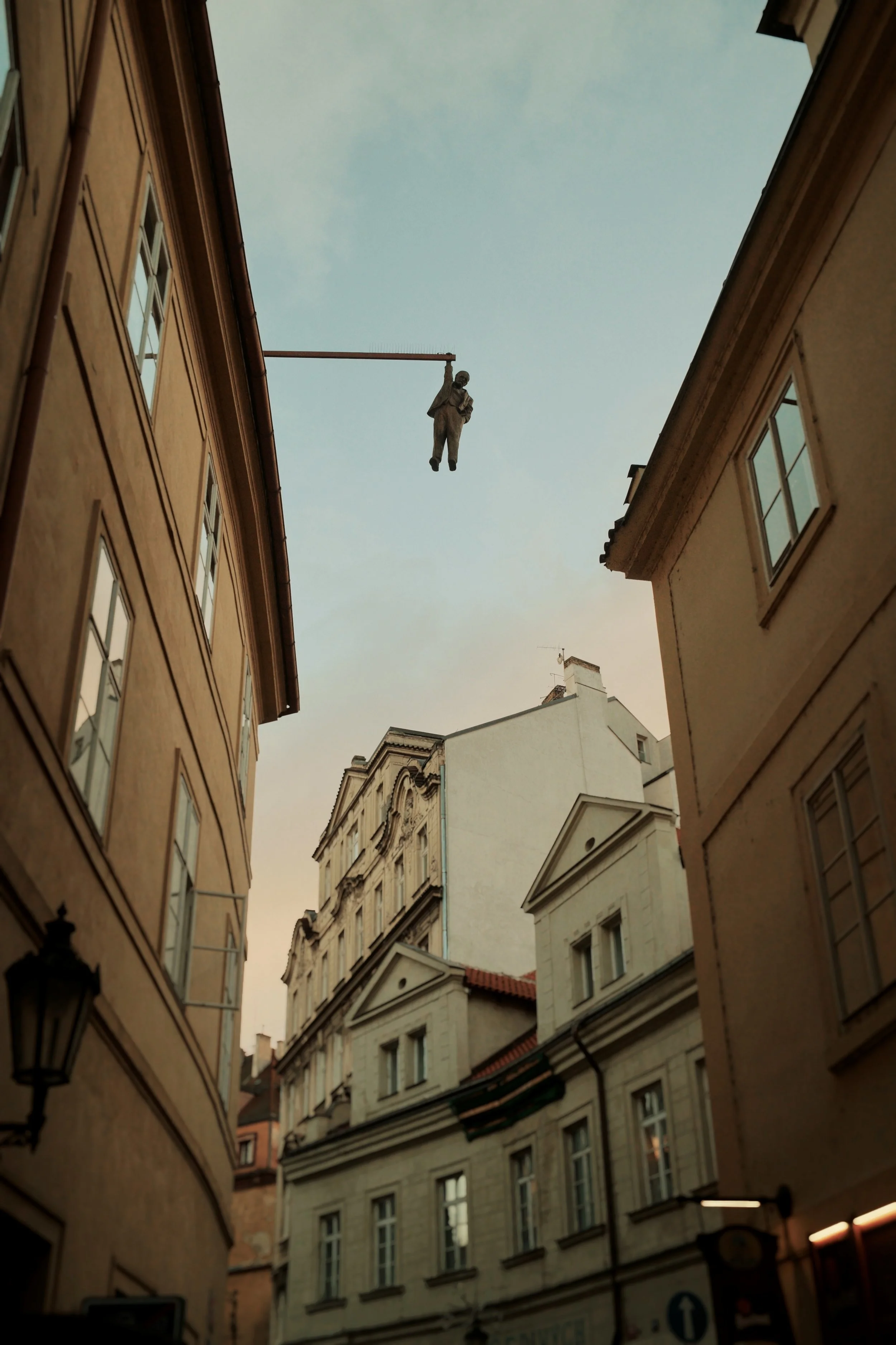 A narrow city alley with European-style buildings on either side, an empty sky visible above, and a sculpture of a person hanging from a wire between the buildings.