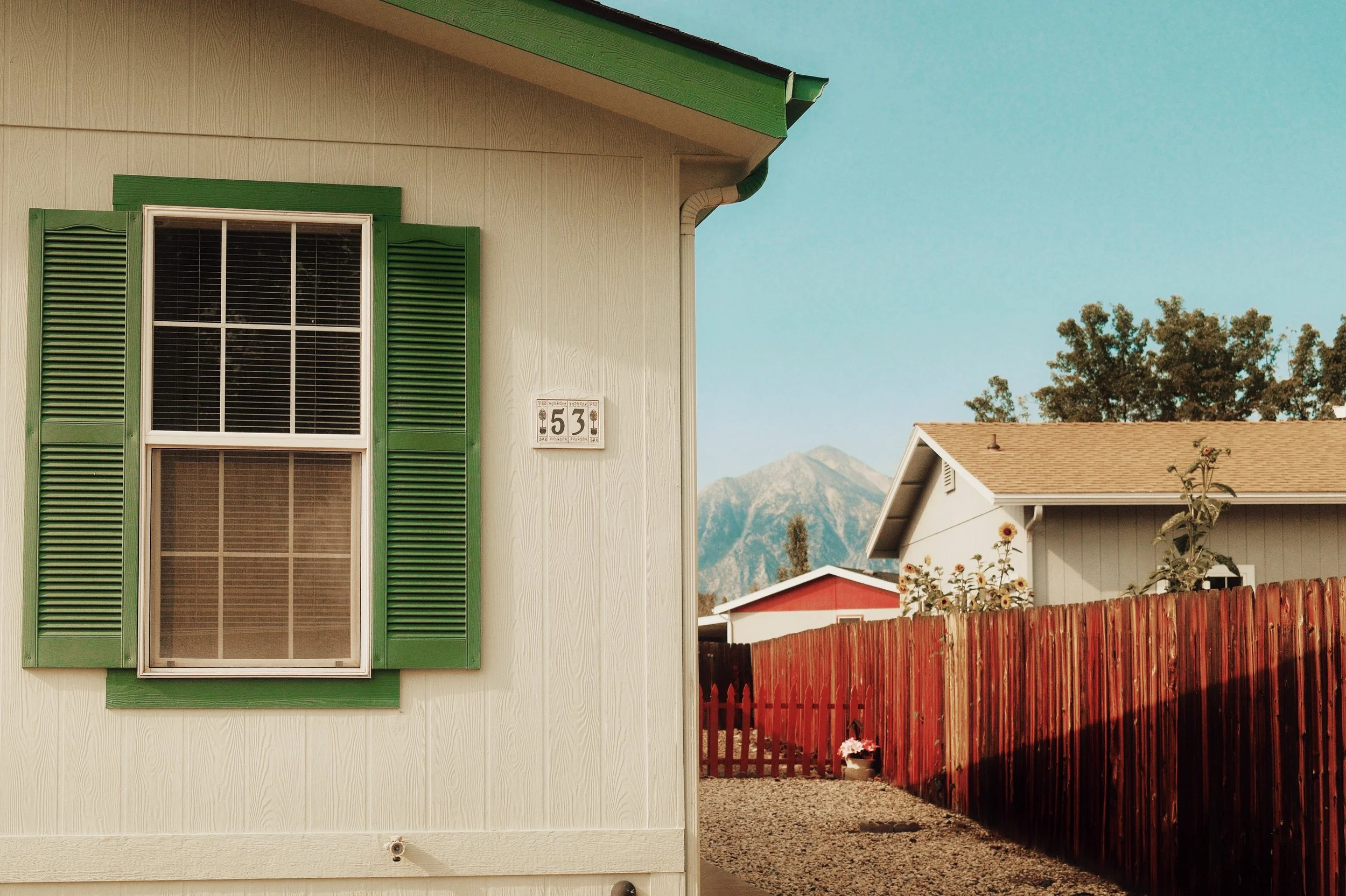 Close-up of a house with green shutters, green siding, and house number 53, with neighbouring houses, a red wooden fence, and mountains in the background.