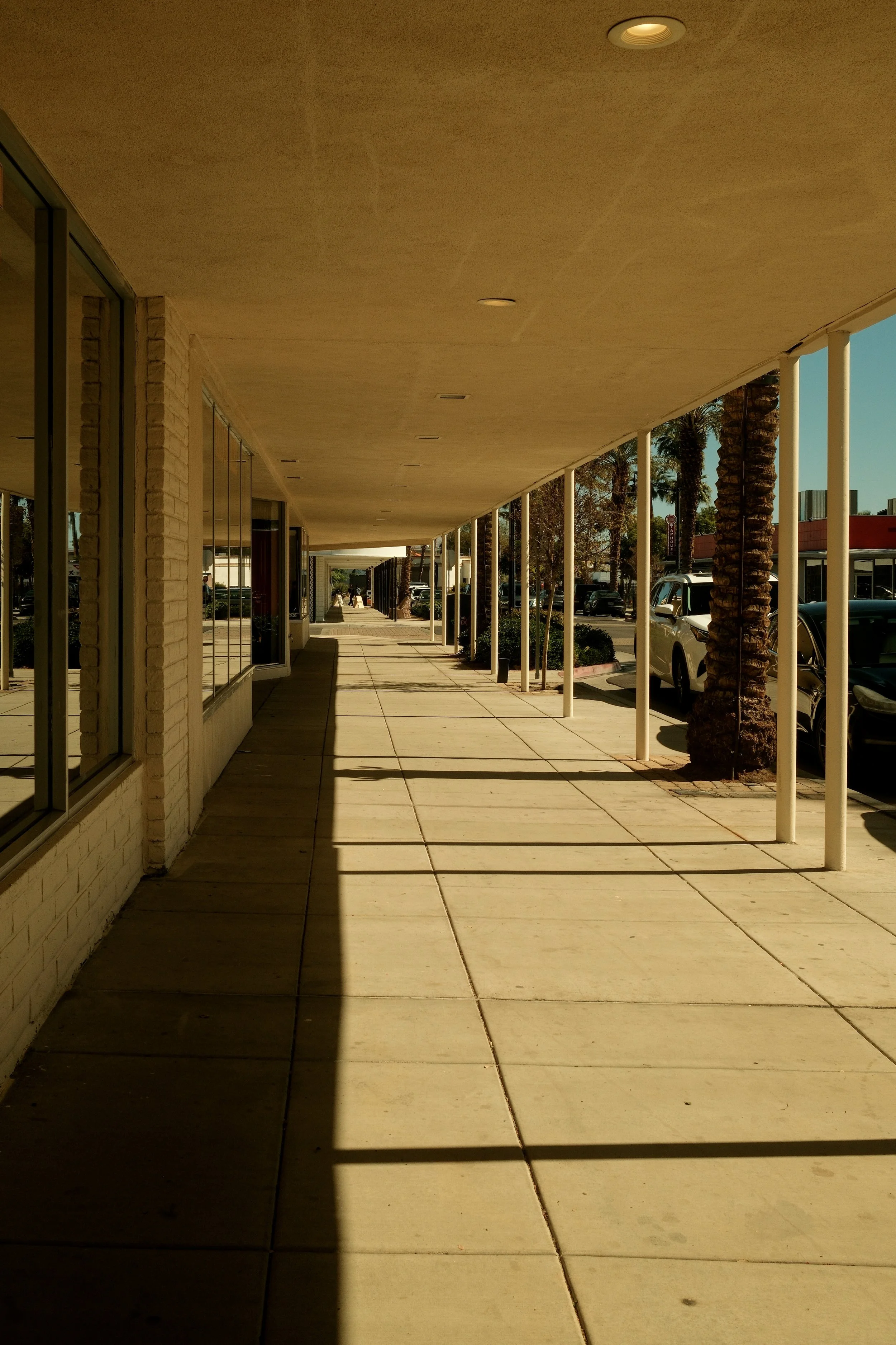 Empty sidewalk in the shade outside a shopping center, with parked cars and palm trees on the right side.