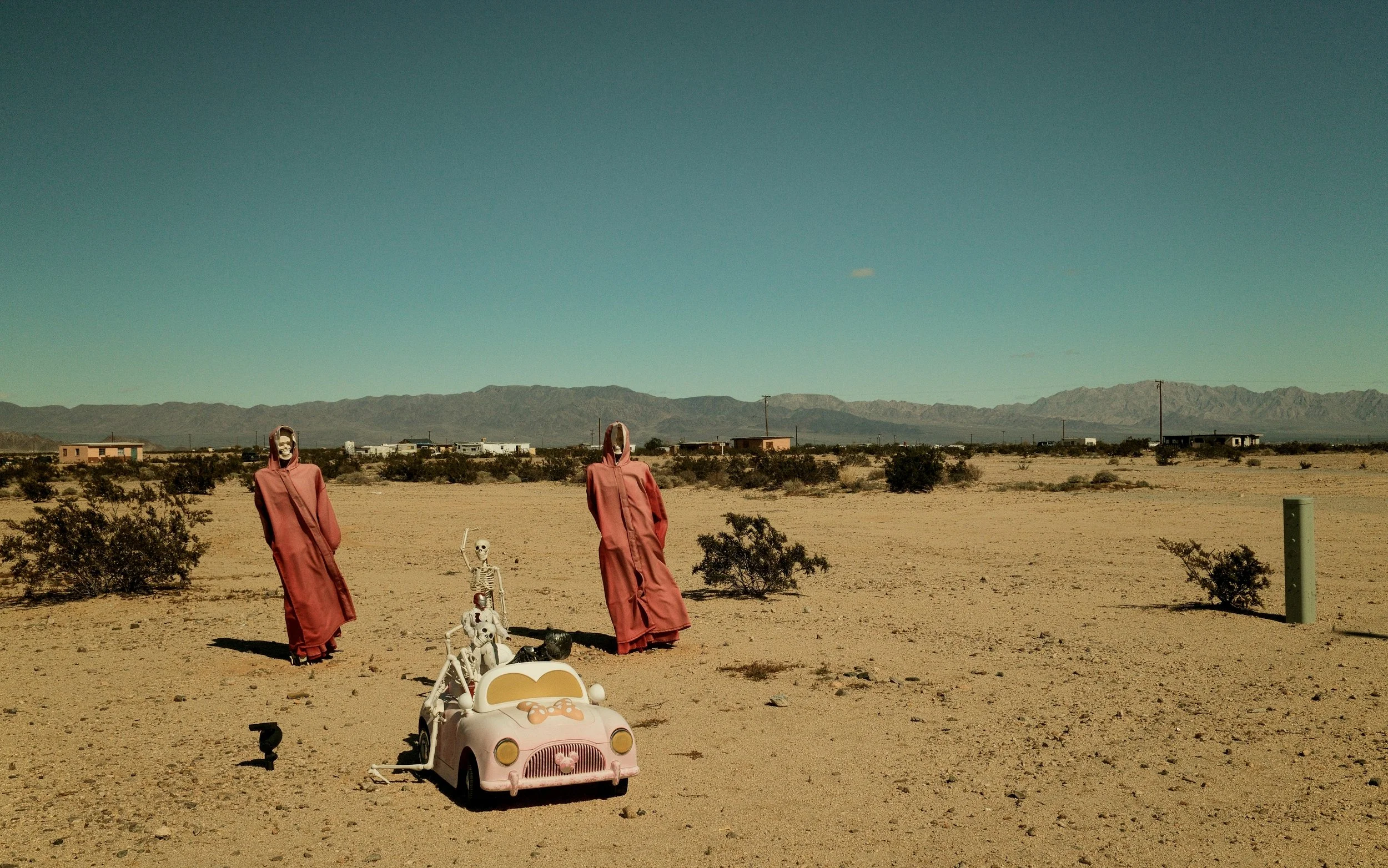 Skeletons dressed in pink robes standing in a desert landscape with a small toy car and a skeleton in the driver's seat.
