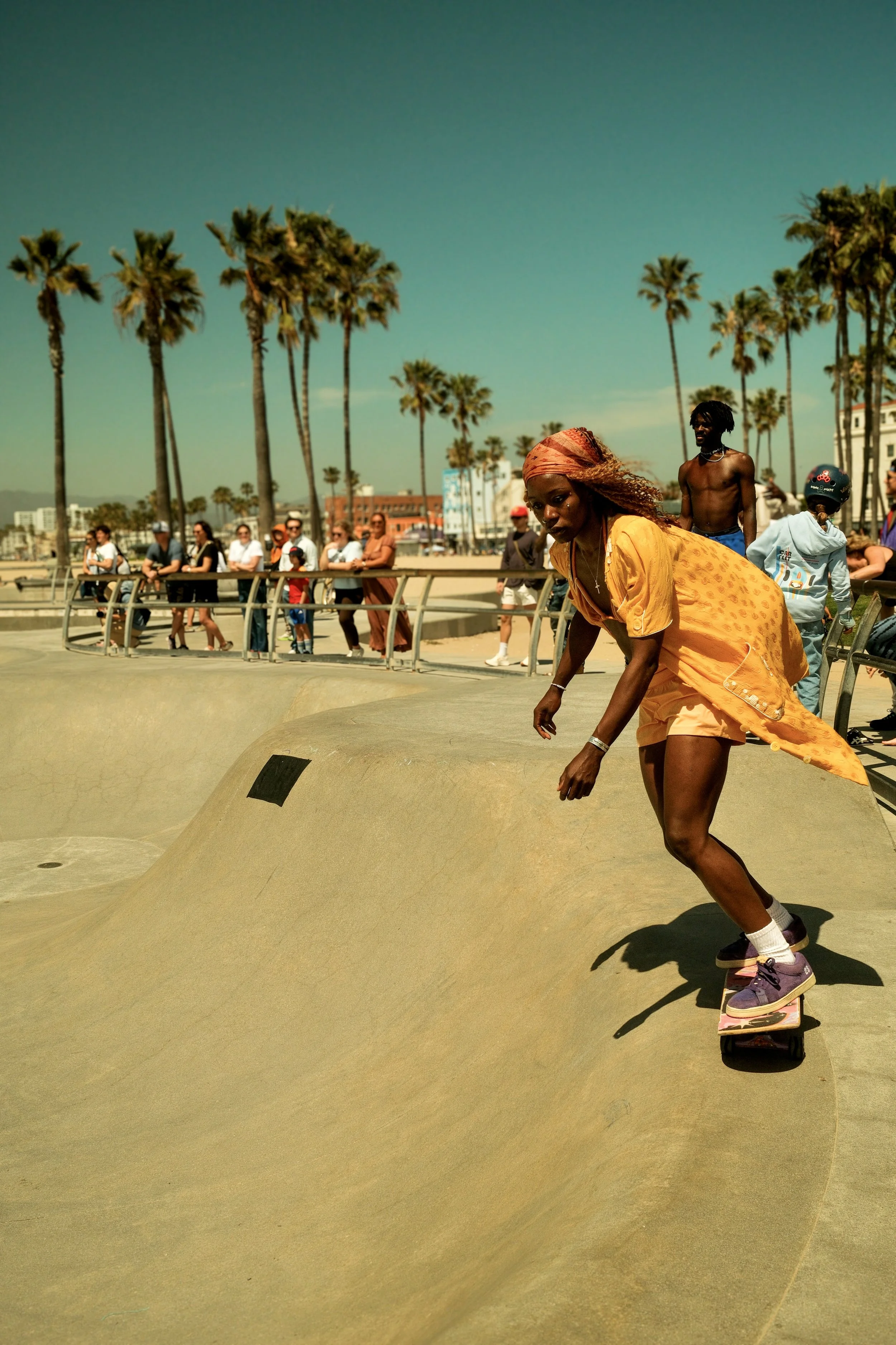 A woman skateboarding at a skate park with palm trees and spectators in the background.