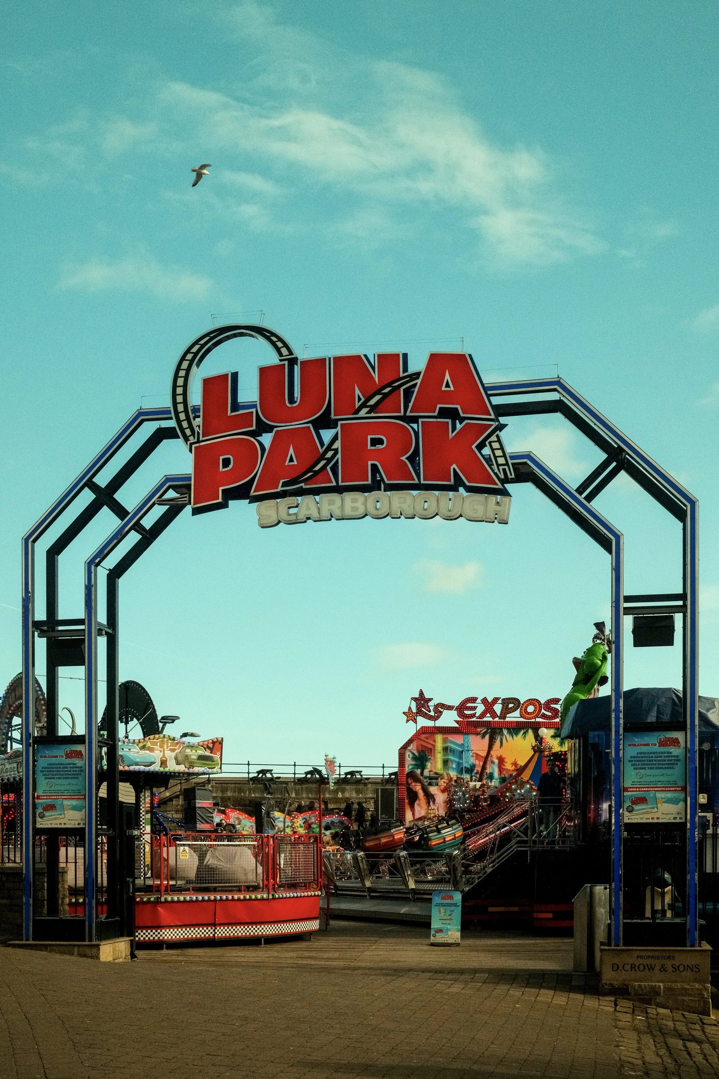 Entrance to Luna Park amusement park in Scarborough, North Yorkshire with rides and attractions, clear sky, a bird flying overhead, and colorful signage.
