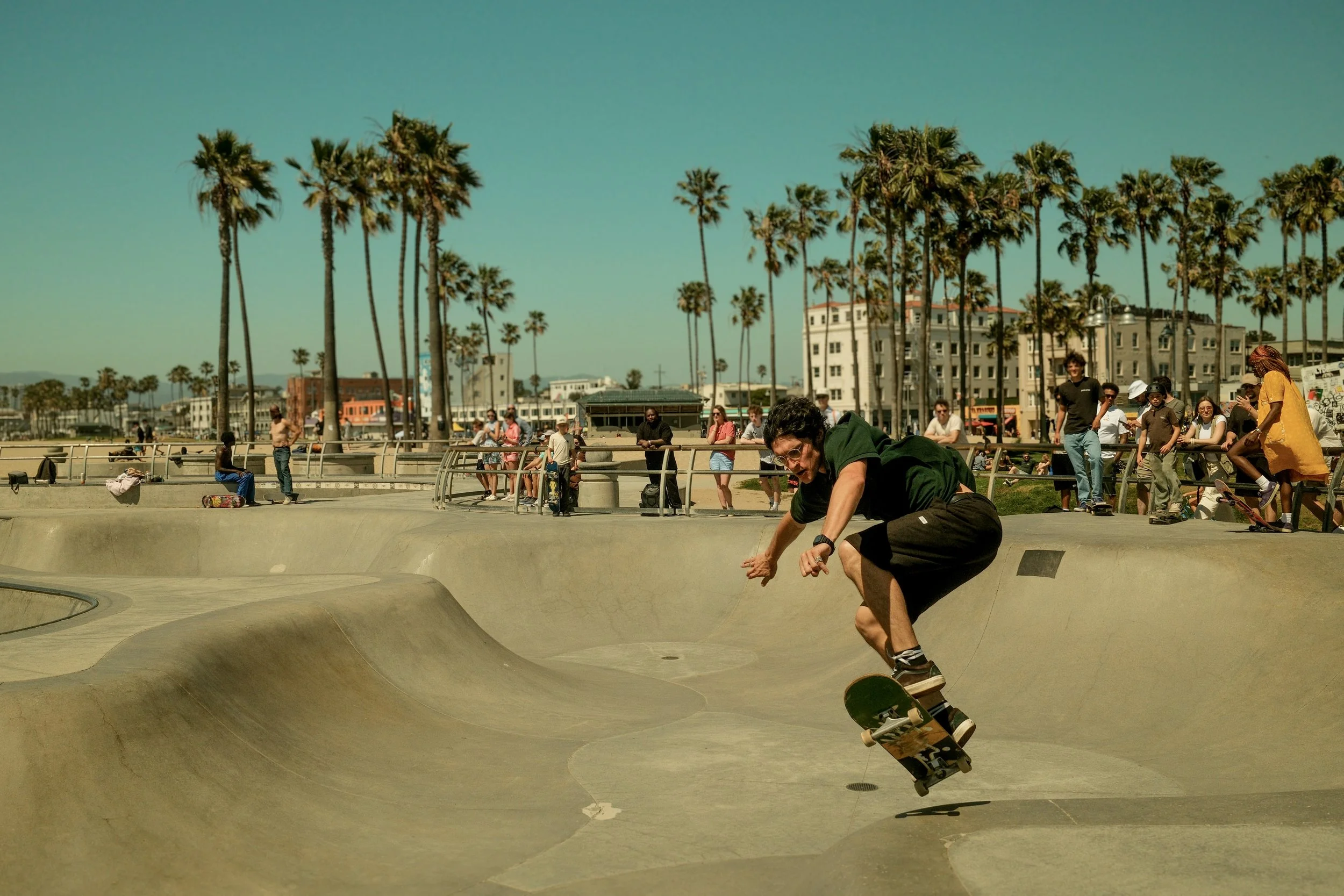 A skateboarder is mid-air performing a trick in a concrete skatepark with an audience and palm trees in the background.