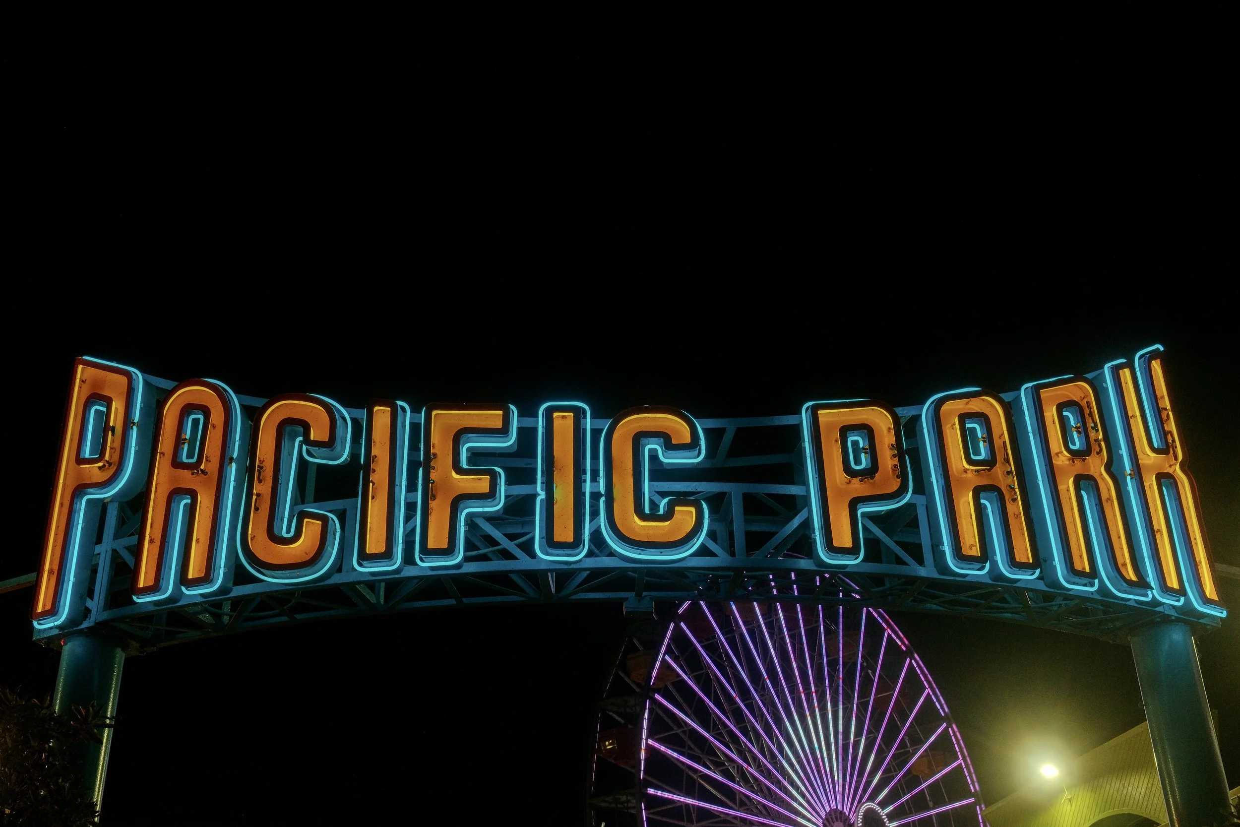 Neon sign reading 'Pacific Park' with a Ferris wheel in the background at night.