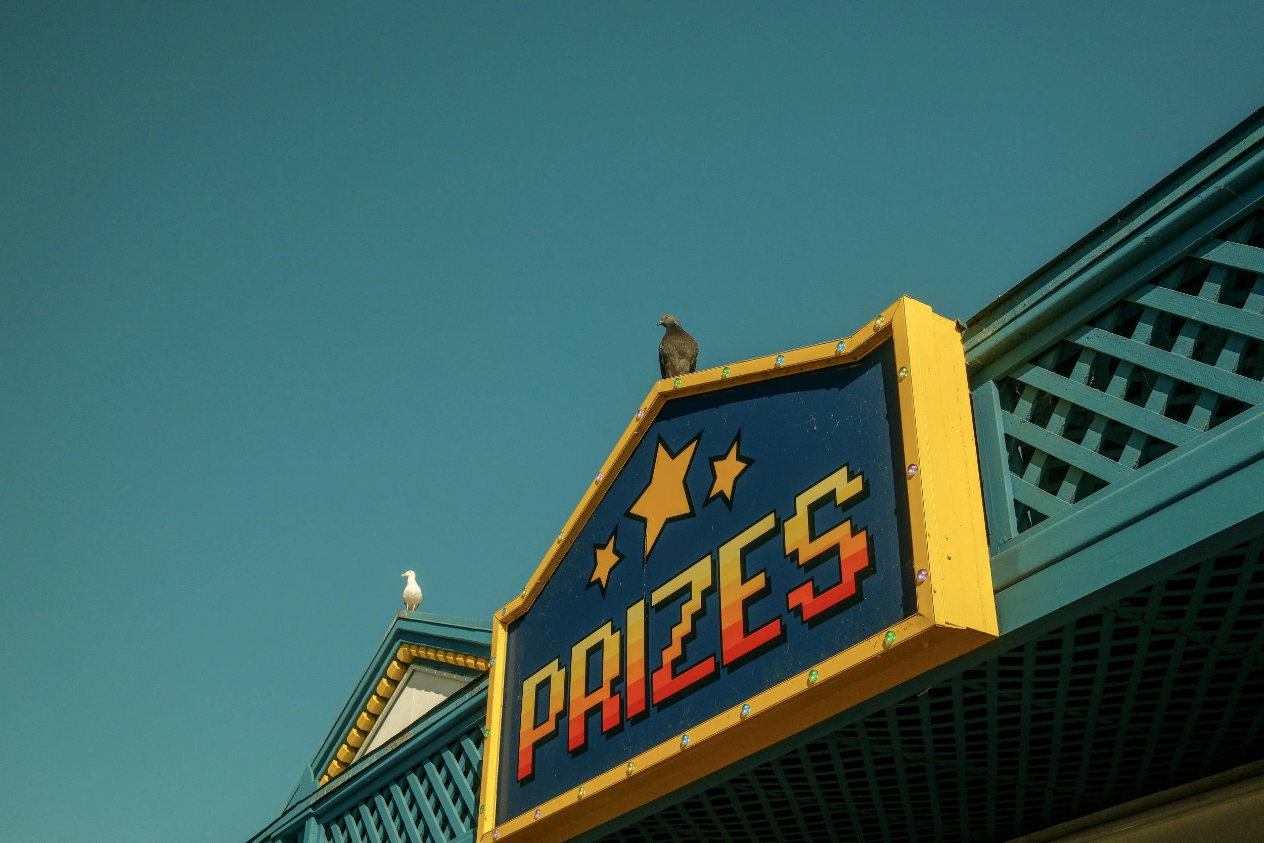 Colorful carnival game booth with a sign that reads 'Prizes' and stars, with two birds perched on top against a clear blue sky.