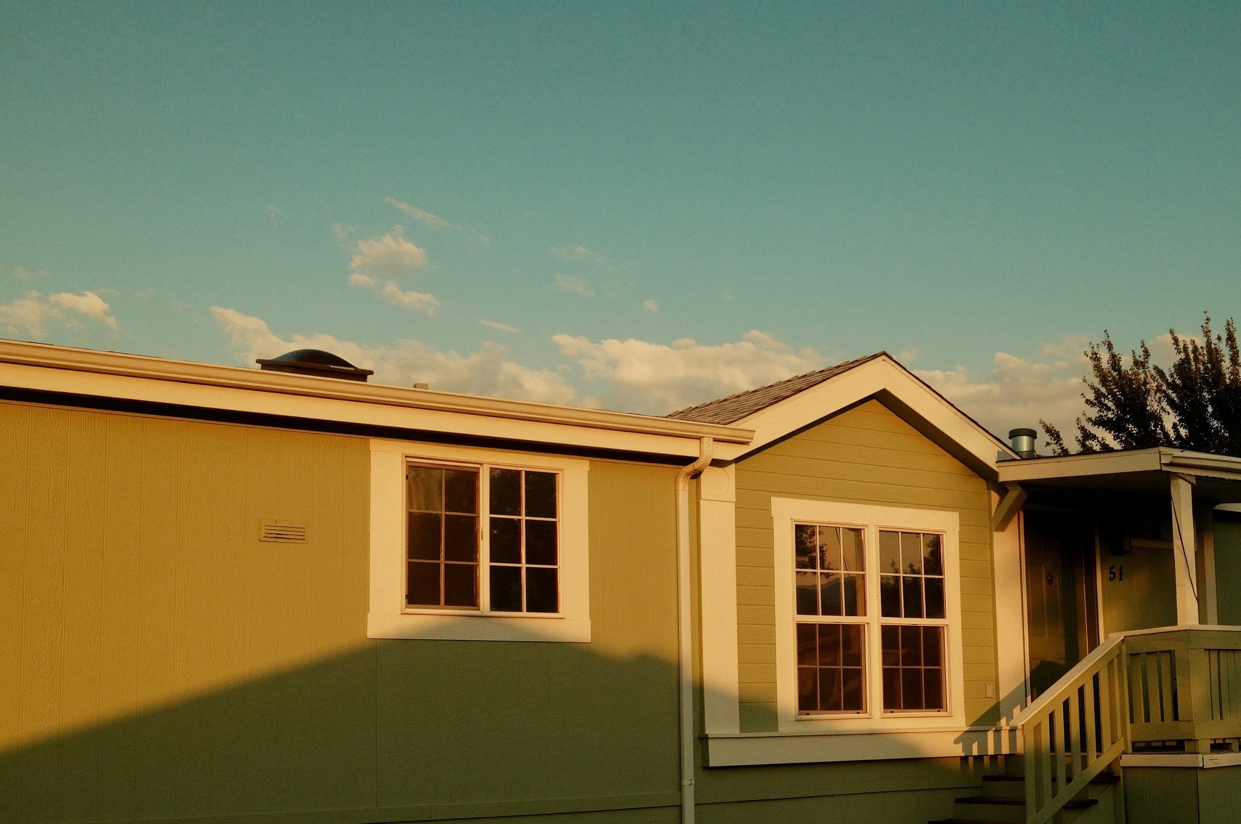 A house painted in light green with white trim, featuring multiple windows, an outdoor staircase, and a roof, with trees and a partly cloudy sky in the background.