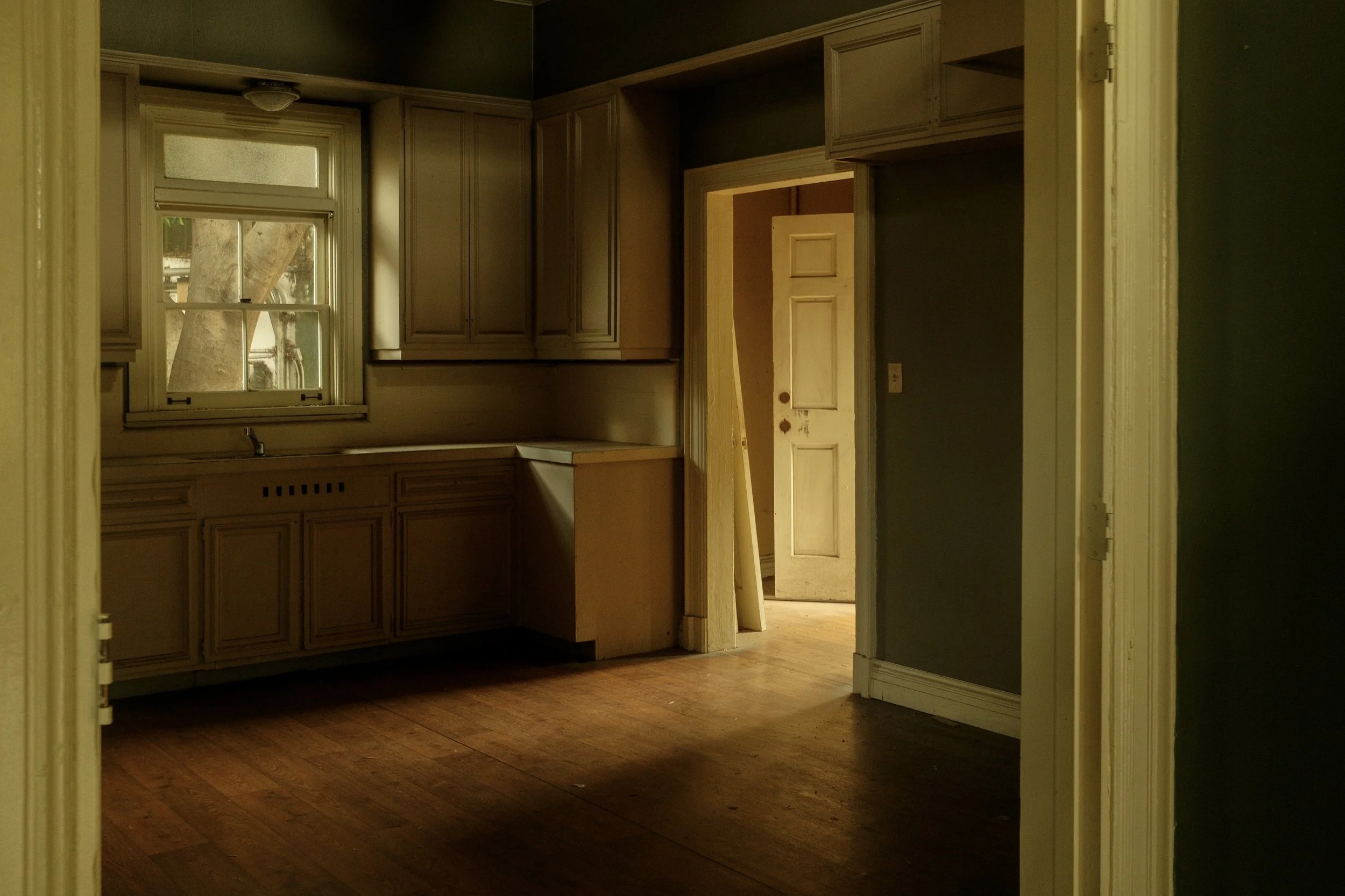 Empty kitchen with wooden floors, beige cabinets, a window, and an open doorway leading to a room with a door.