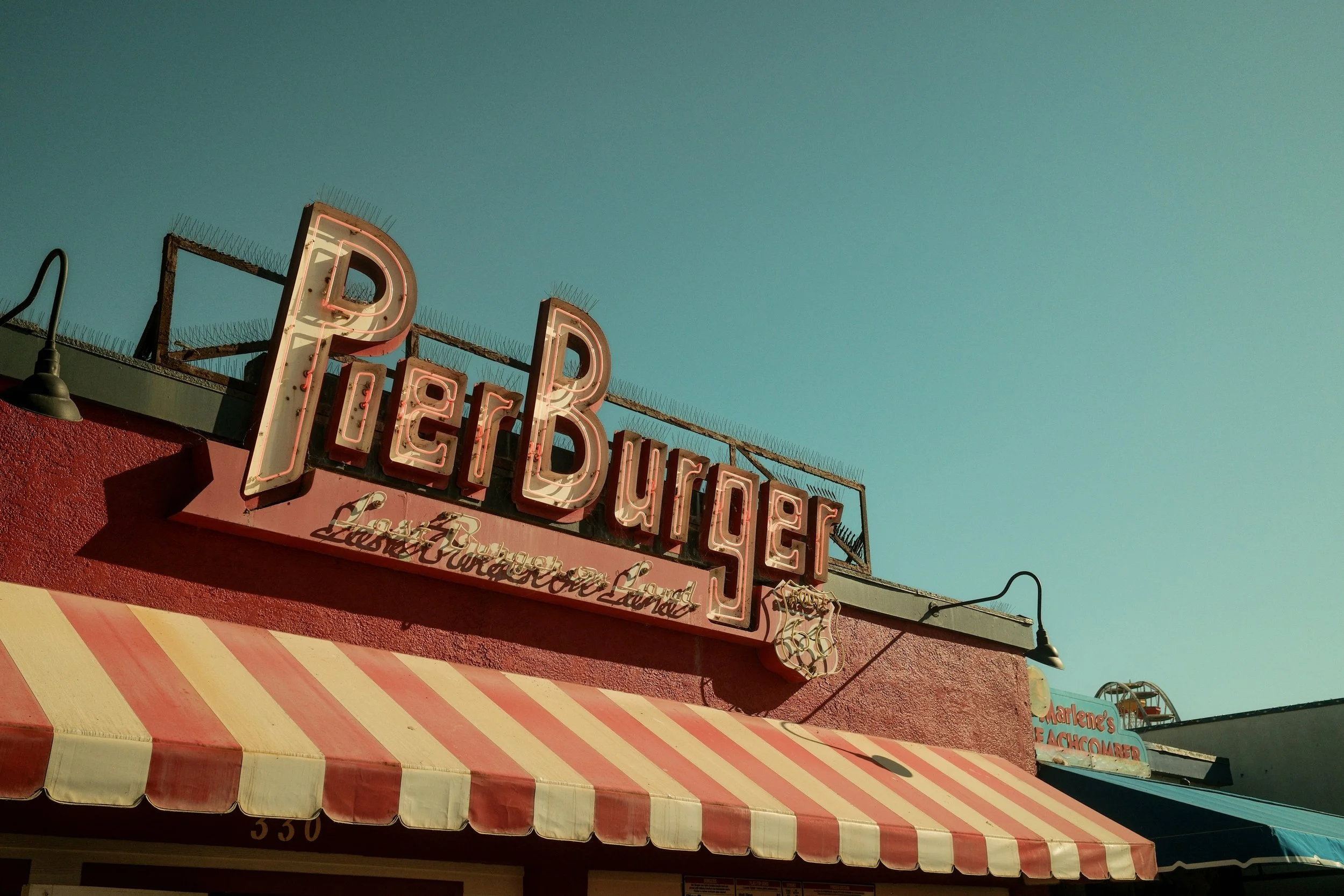Neon sign for Pier Burger restaurant on a pink building with a striped red and white awning.