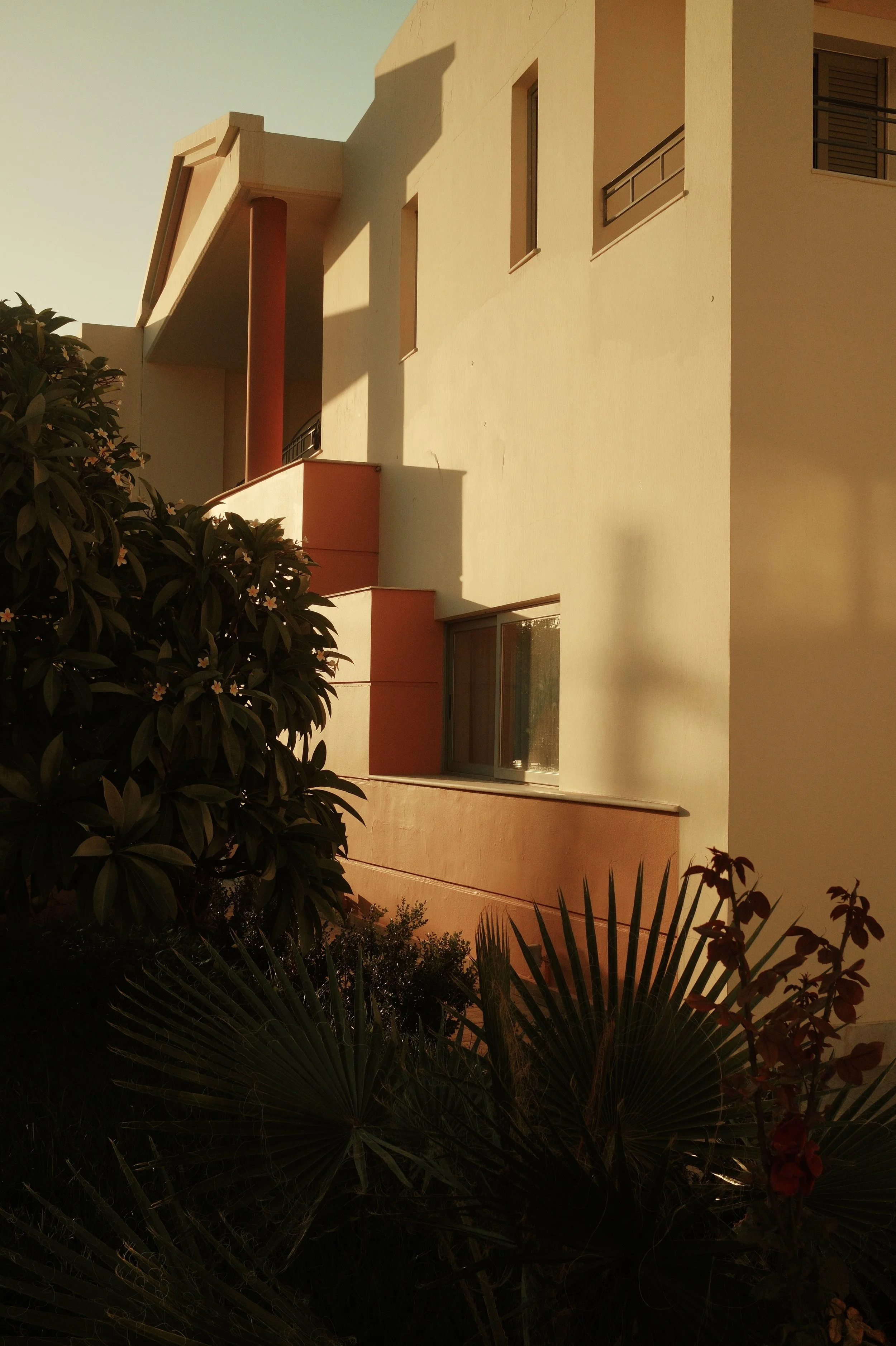 A modern multi-story building with beige and orange exterior walls, small window, and shadowed sections. There are bushes and plants in the foreground.