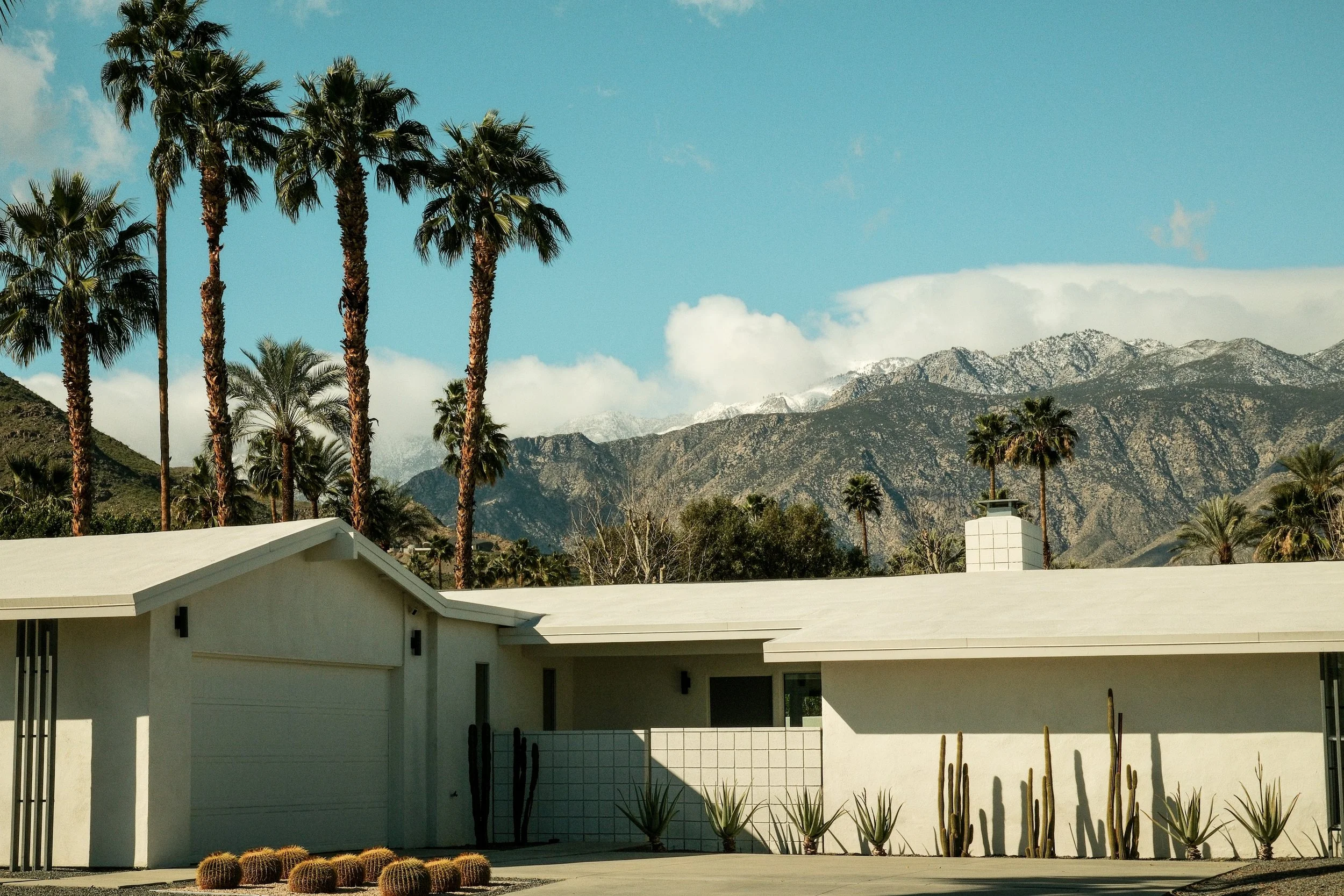 Modern house with white walls and a flat roof, surrounded by tall palm trees and cacti, with mountains in the background under a blue sky with some clouds.