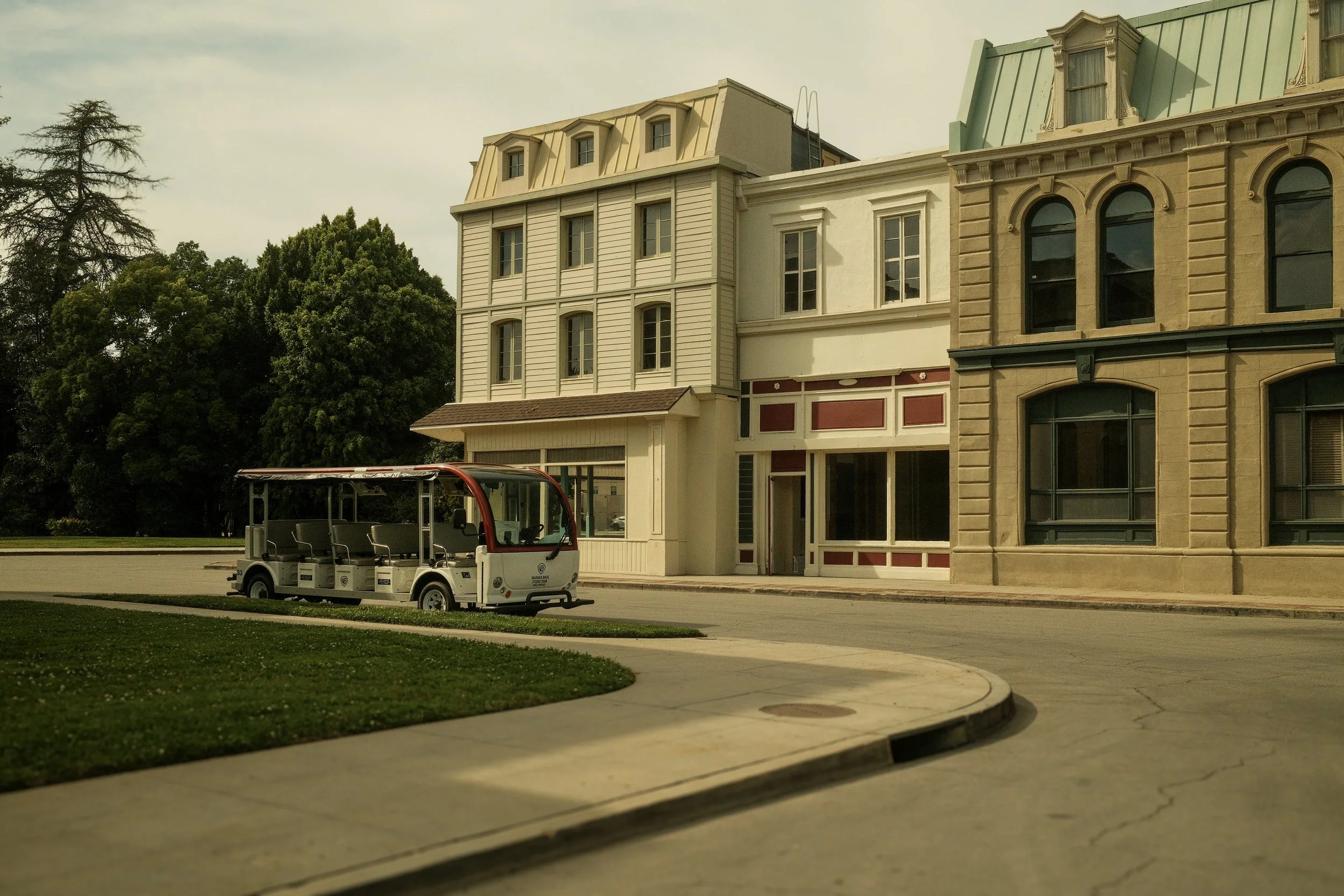 A small open-air shuttle parked on the street in front of historic buildings with large windows and decorative facades, with trees in the background.