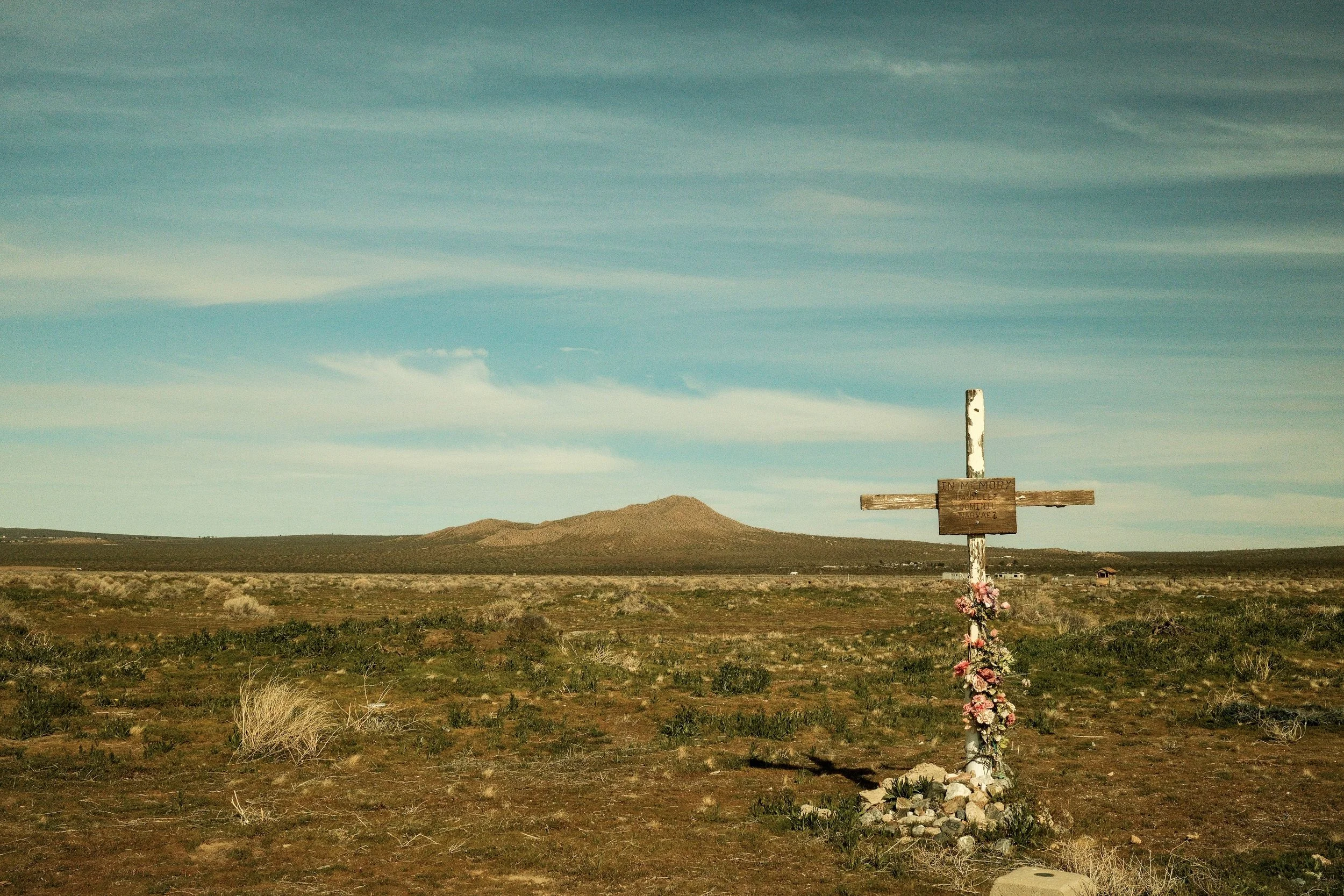 A lone wooden cross with a sign attached stands in a barren landscape, decorated with flowers at its base. In the background, a mountain and a wide open sky with scattered clouds are visible.