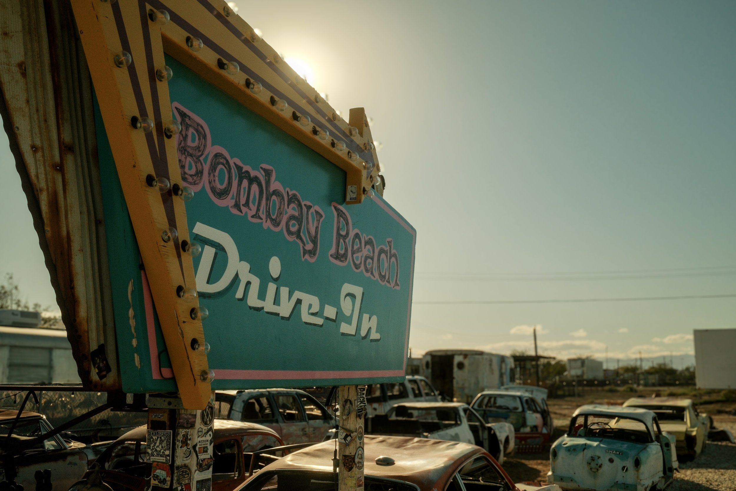 A vintage sign with fluorescent bulbs advertising 'Bombay Beach Drive-In', positioned over a lot of abandoned, rusted cars with a desert landscape and clear sky in the background.