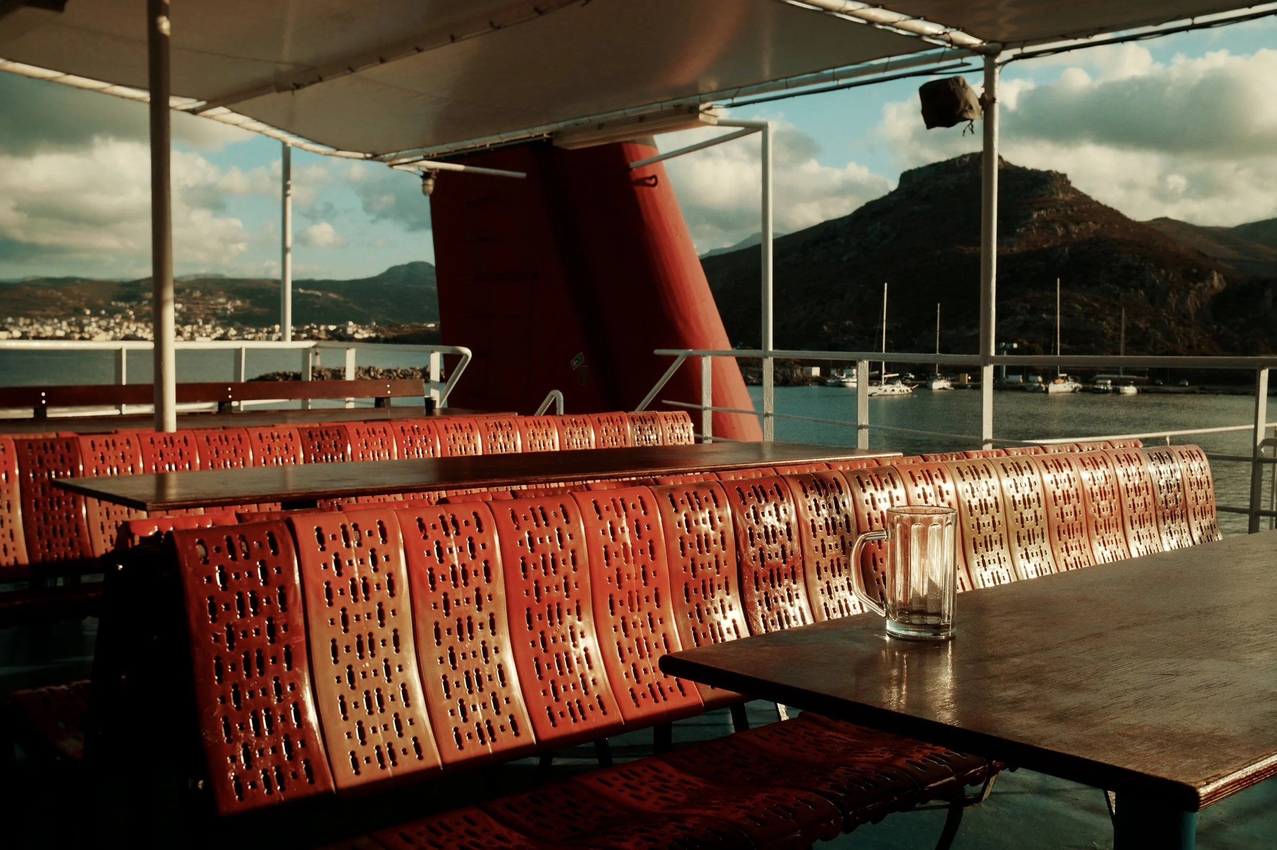 Empty outdoor deck with red chairs, a wooden table with a glass mug, overlooking a harbor with boats and mountains in the distance, under a partly cloudy sky.