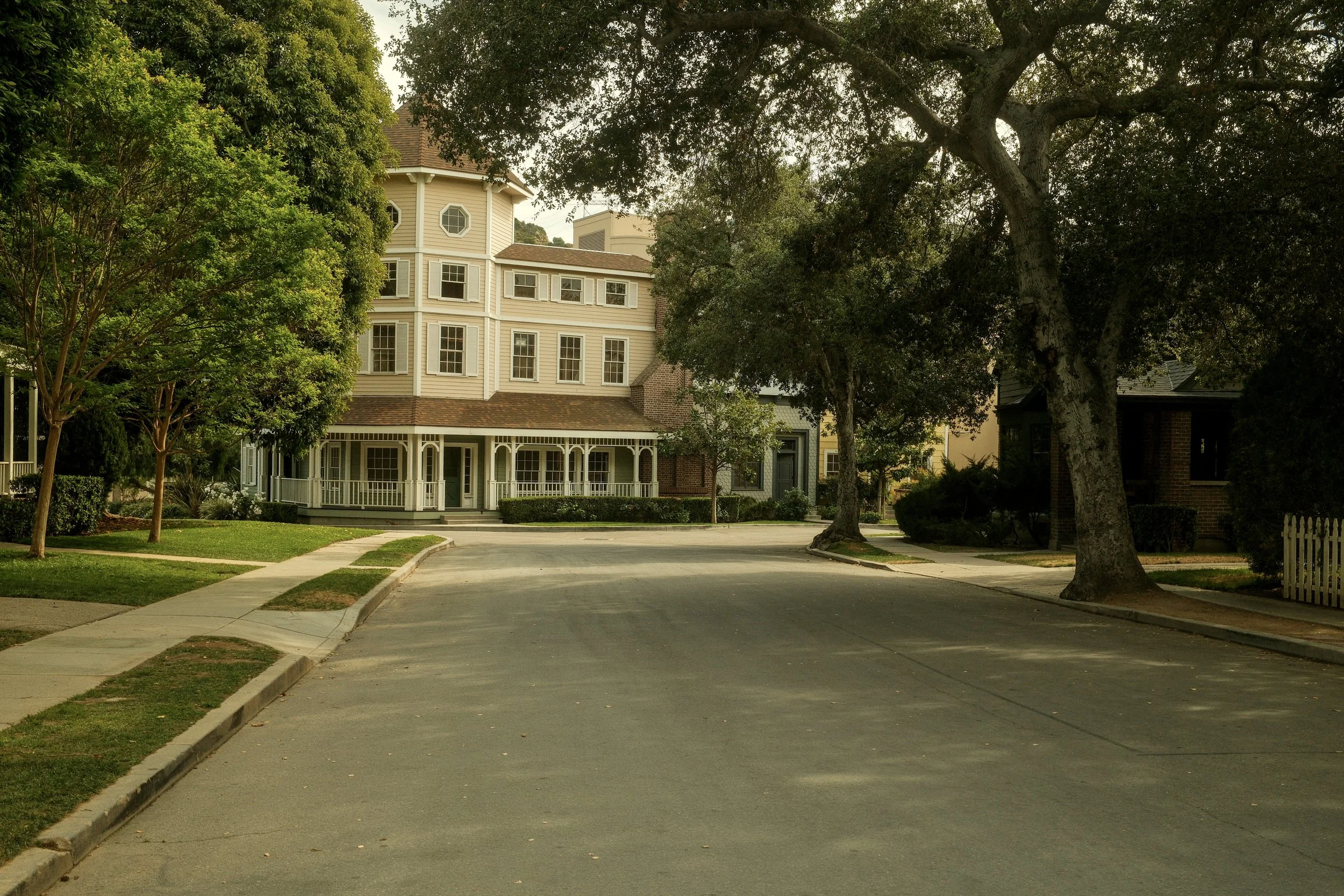 Residential street scene with a large beige multi-story house, trees, and sidewalks.