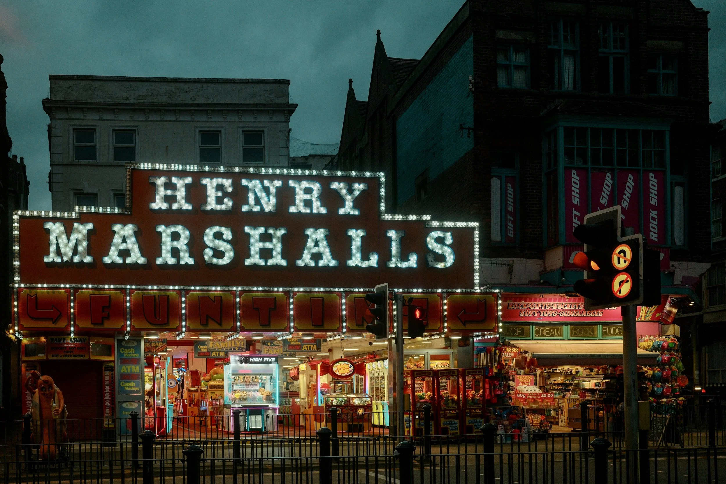 A colorful city attraction with a large illuminated sign reading 'Henry Marshalls' in front of food stalls and game booths, with buildings and a darkening sky in the background.