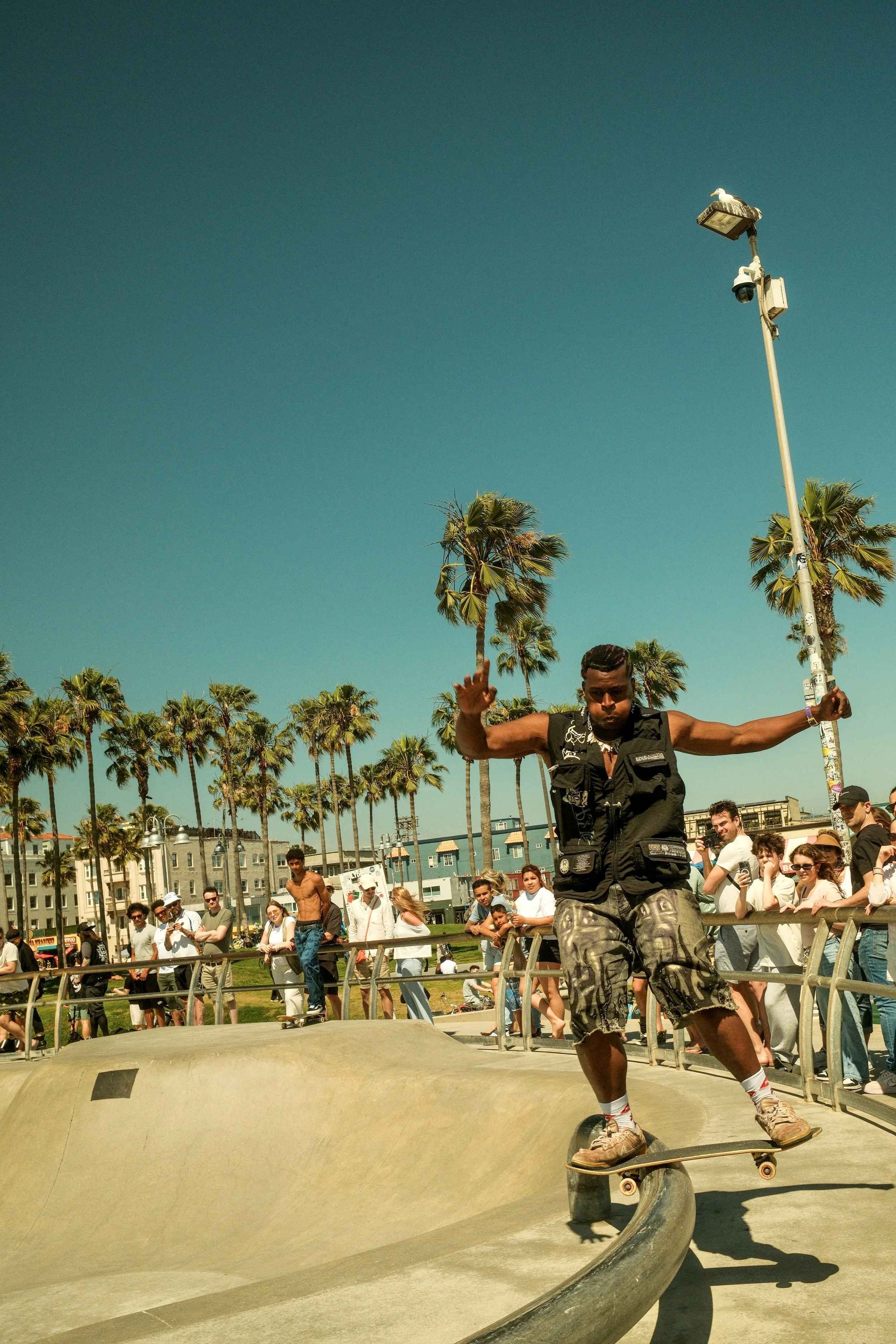Skateboarder performing a trick at a skate park with a crowd of spectators, palm trees, and a clear blue sky in the background.