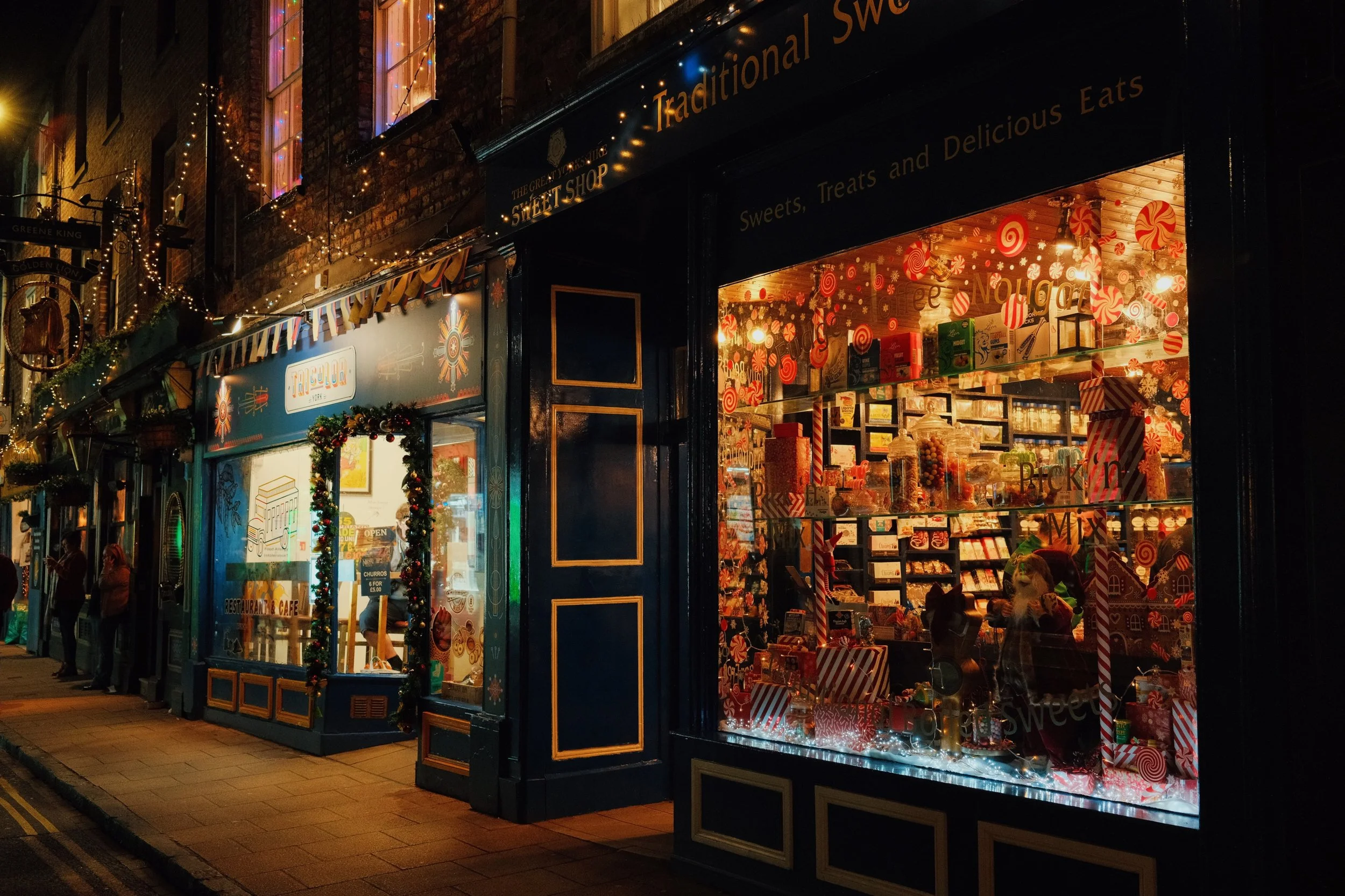 A decorated Christmas shop window with holiday treats, candy cane displays, and festive lights, on a city street at night.