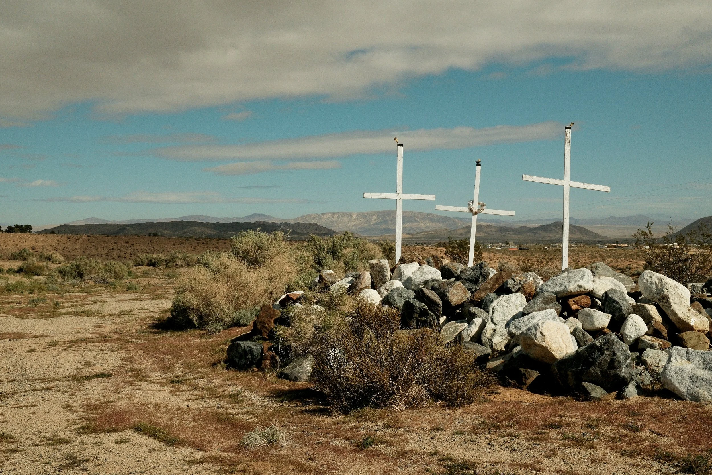 Three white crosses mounted on rocks in a desert landscape with mountains in the distance and a partly cloudy sky.
