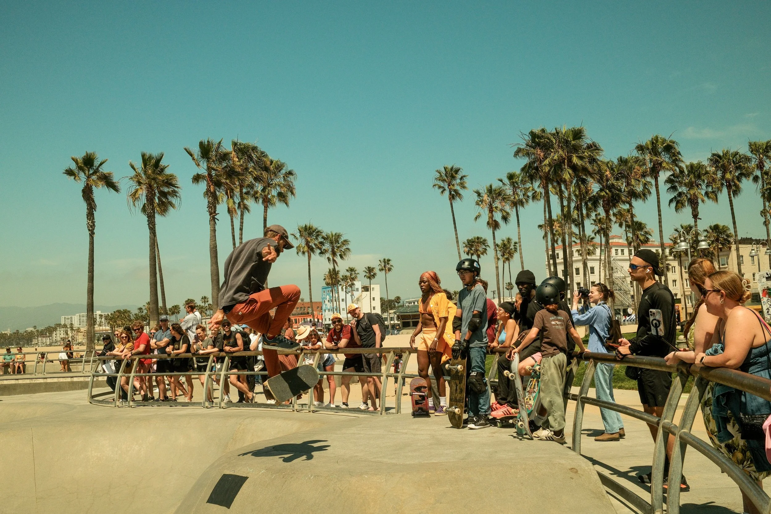Skateboarder jumps over rail at a busy skatepark with palm trees and buildings in the background, and spectators watching.