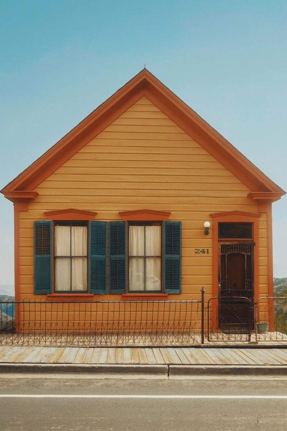 A yellow wooden house with blue shutters, a small front porch, and a black metal fence, located by a paved road, under a clear sky.