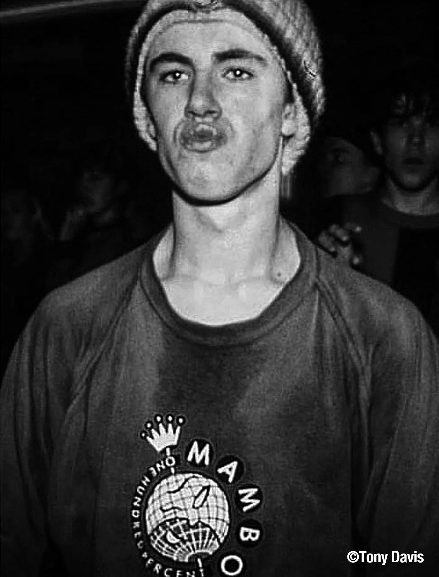 Black and white photo of a young man wearing a beanie and a T-shirt with a globe and crown graphic, making a duck face.