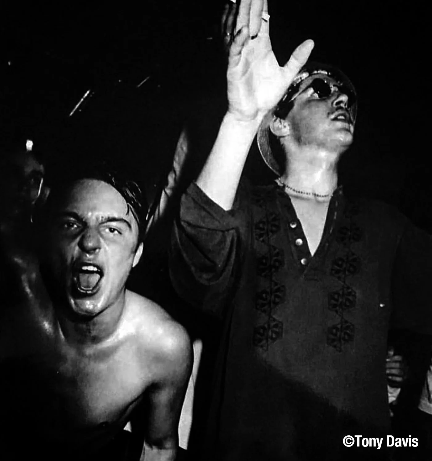 Black and white photo of two young men. The shirtless man on the left appears to be shouting, with his mouth open and arm raised. The man on the right is wearing sunglasses, a cap, and a dark shirt with embroidery, raising his hand.
