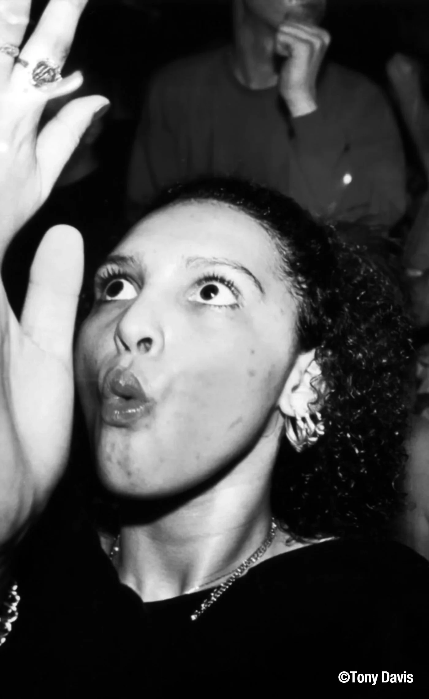 A woman with curly hair making a playful face with pursed lips and wide eyes, holding her hand near her face, woman wearing earrings and a necklace.